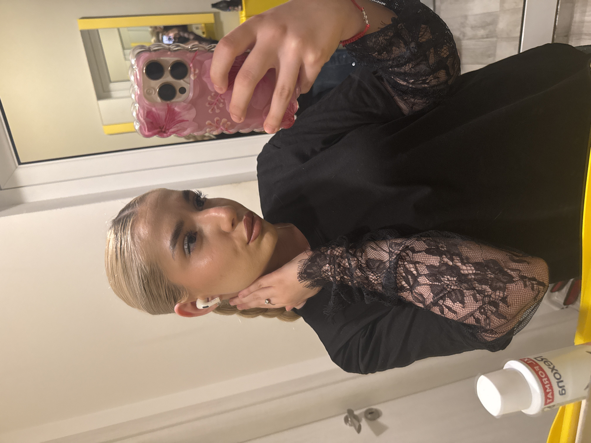 A young woman with blonde hair, styled back from her face, is taking a mirror selfie indoors. She is looking directly into the smartphone's camera, which is held by her right hand. The phone has a distinct pink, floral-patterned case. A white earbud is visible in her left ear, and a silver ring with a clear stone is on her left ring finger. On her right wrist, a red string bracelet is visible.
She is wearing a black top or dress featuring prominent black lace sleeves, adding a decorative element to her attire. The setting appears to be an indoor room with light-colored walls, reflected in the mirror. A yellow frame or shelf is also reflected in the upper left background of the mirror.
In the bottom right corner of the image, partially obscured and reflected in the mirror, is a white bottle with red lettering. The visible, reversed text on the bottle appears to read "SKORUEK," suggesting the brand name "KEROUS" or similar when viewed correctly. The overall activity is a self-portrait taken in an indoor environment, possibly in Sofia, Bulgaria, though no specific visual cues for the location are present in the image itself.