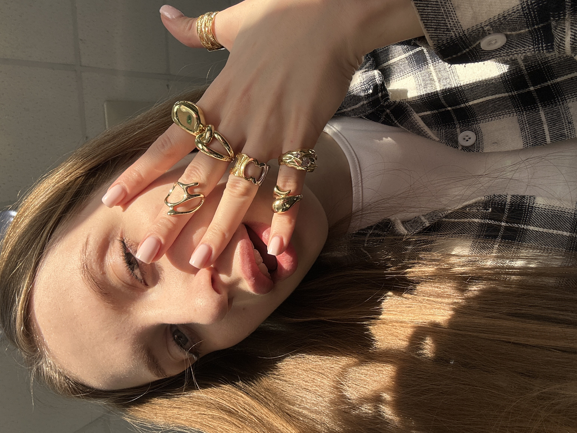 An angled, close-up indoor portrait depicts an individual in the city of Chaikovsky, Russia. The subject, appearing to be a young person with long, light brown hair and fair skin, is positioned to the right of the frame, looking towards the camera. They are wearing a white undershirt beneath an open black and white plaid flannel shirt. The subject's left hand is raised, with fingers partially obscuring the lower portion of the face, specifically the mouth and nose area. The visible fingernails are manicured with a light pink polish. This hand is heavily adorned with multiple gold-colored rings on most fingers, including abstract, knotted, heart-shaped, and textured band designs. Strong natural light illuminates the subject from the left, creating bright highlights on the hair and face, and casting distinct shadows. The background is out of focus, revealing light-colored ceiling tiles with visible grid lines, indicative of an indoor setting.