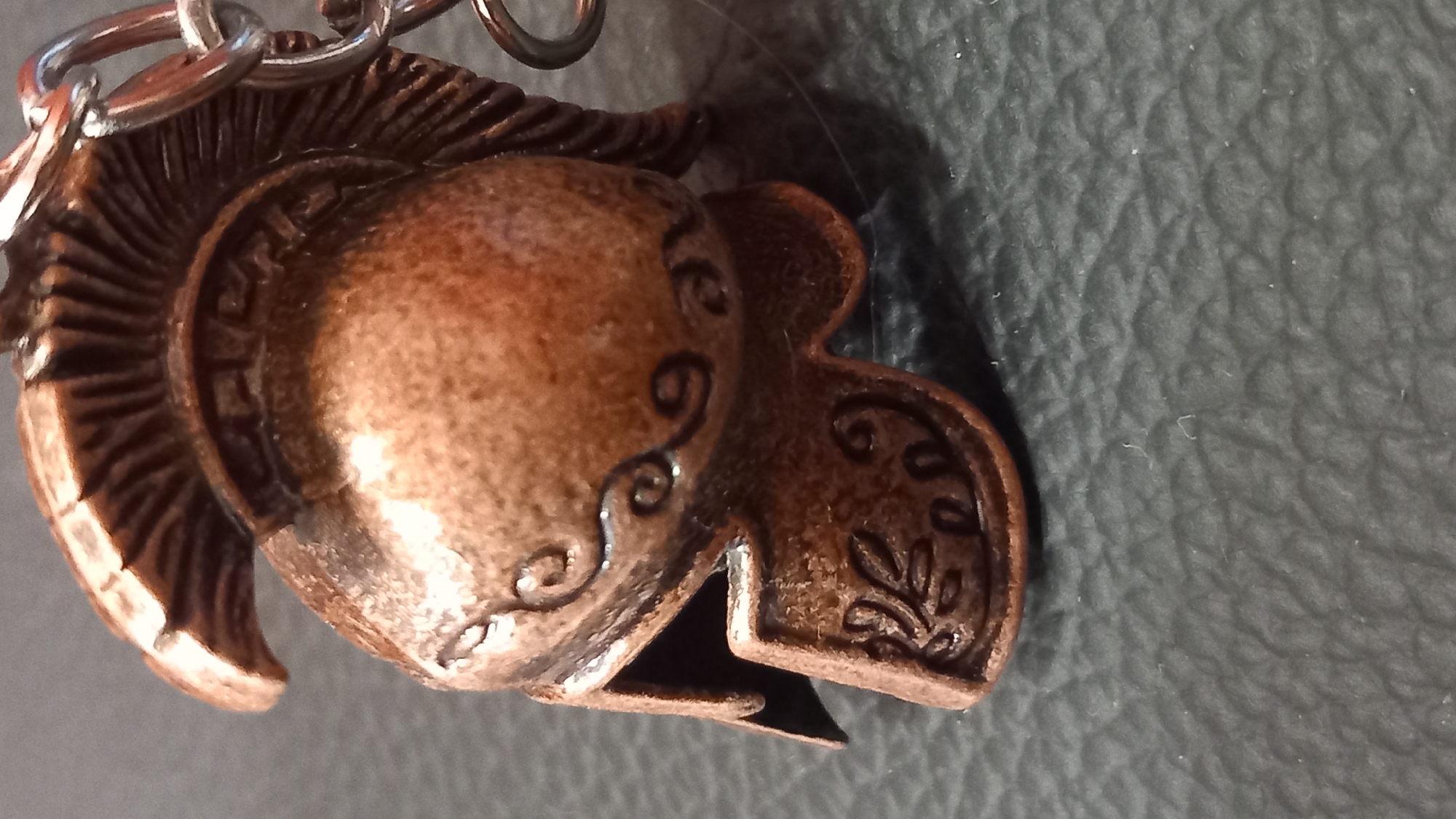 The image is a close-up, shallow depth of field shot of a miniature bronze spartan helmet, likely a keychain or decorative item. The helmet is detailed with embossed swirling patterns and a laurel leaf design on the cheek guard. It has a stylized crest of vertical ridges. A silver-colored chain loop is attached to the top right of the helmet. The helmet is resting on a dark, textured surface, possibly leather or a similar material. The background is out of focus. There are no people or actions depicted. The location in the city of Kos, Greece, is not discernible from the image.