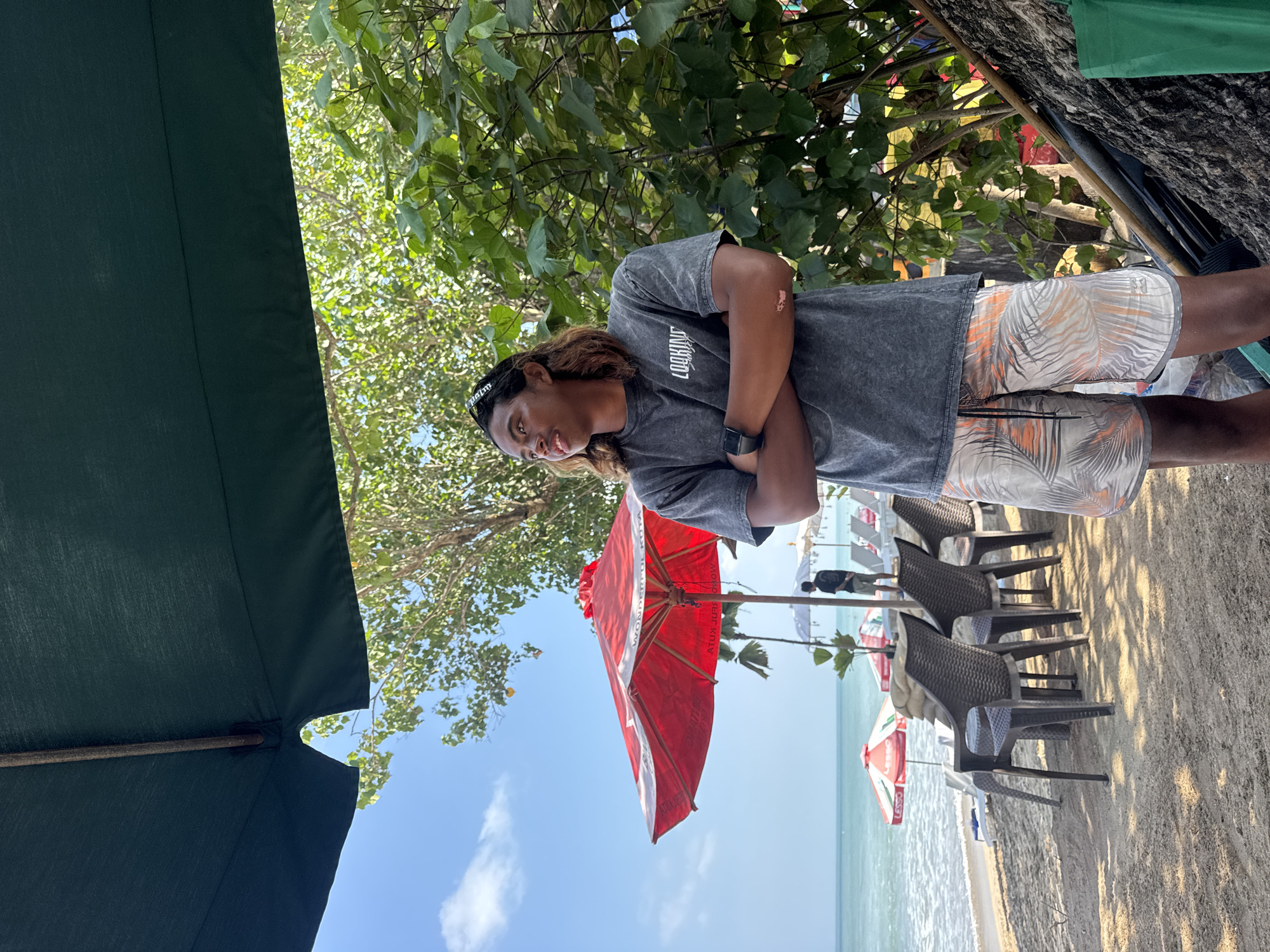 A young man stands on a sandy beach in Kuta, Indonesia, with his arms crossed and looking to his left. He is wearing a dark grey t-shirt with a white graphic and patterned shorts. Behind him, beach chairs and red and white umbrellas are visible, leading to the ocean. Lush green trees and foliage are to his right, and a large dark umbrella shades the foreground. The sky is blue with scattered white clouds, indicating a bright, sunny day.