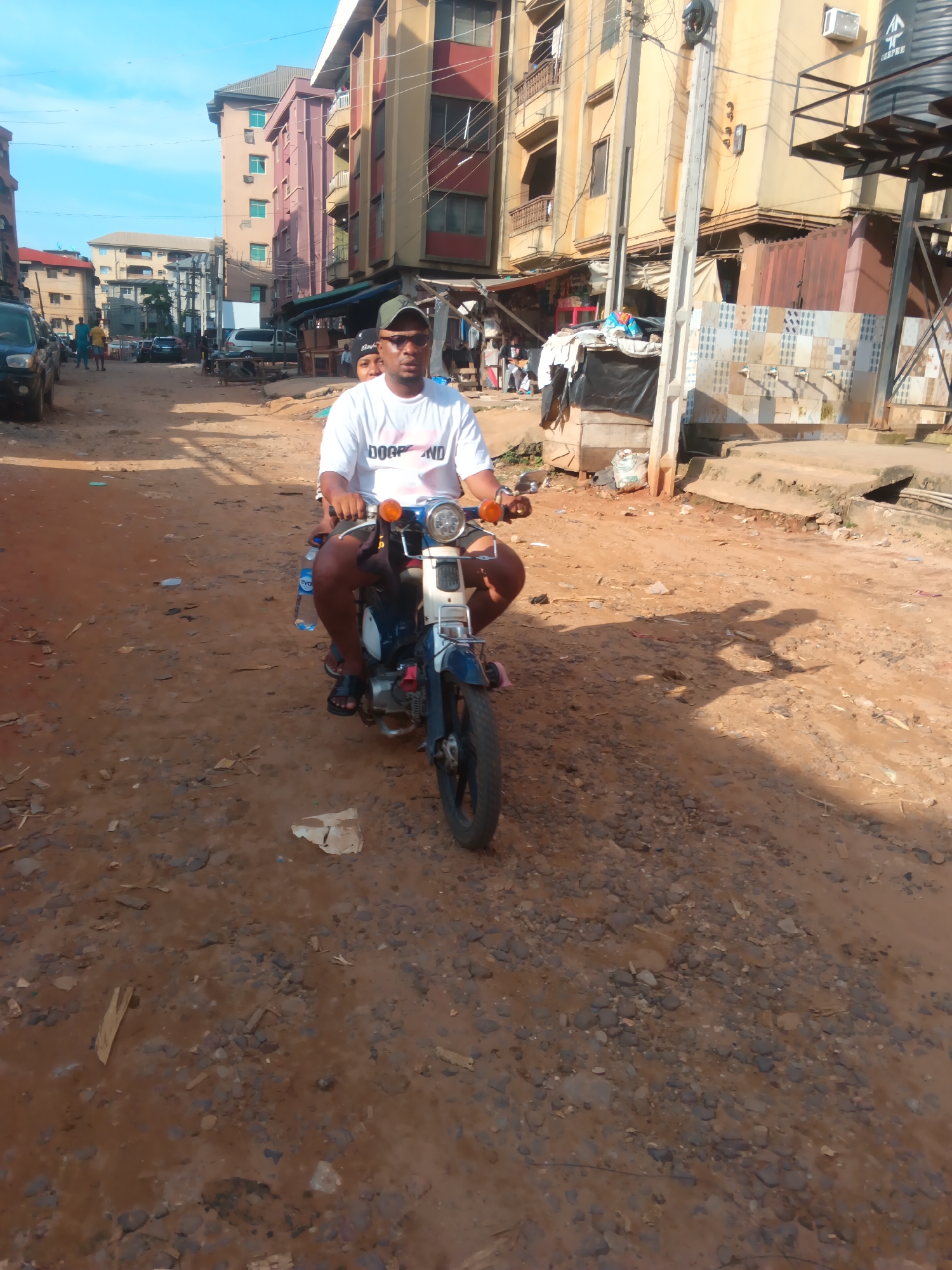 A man and a child are riding on a motorcycle down a dirt road in Onitsha, Nigeria. The man is wearing a white t-shirt with the text "DOOP ND" visible on it, a green baseball cap, and sunglasses. He is holding the handlebars of the blue and white motorcycle. The child is sitting behind him, partially obscured by his body. The road is unpaved and covered in dirt and gravel, with some scattered debris. In the background, there are multi-story buildings lining the street, suggesting an urban environment. One building appears to be a residential apartment complex with balconies, while another has a more commercial feel with stalls and awnings. Power lines and poles are visible, along with a large water tank. The weather appears to be clear and sunny, with a bright blue sky. The time of day is likely daytime, given the lighting and shadows. The overall scene depicts everyday life in a busy Nigerian city.