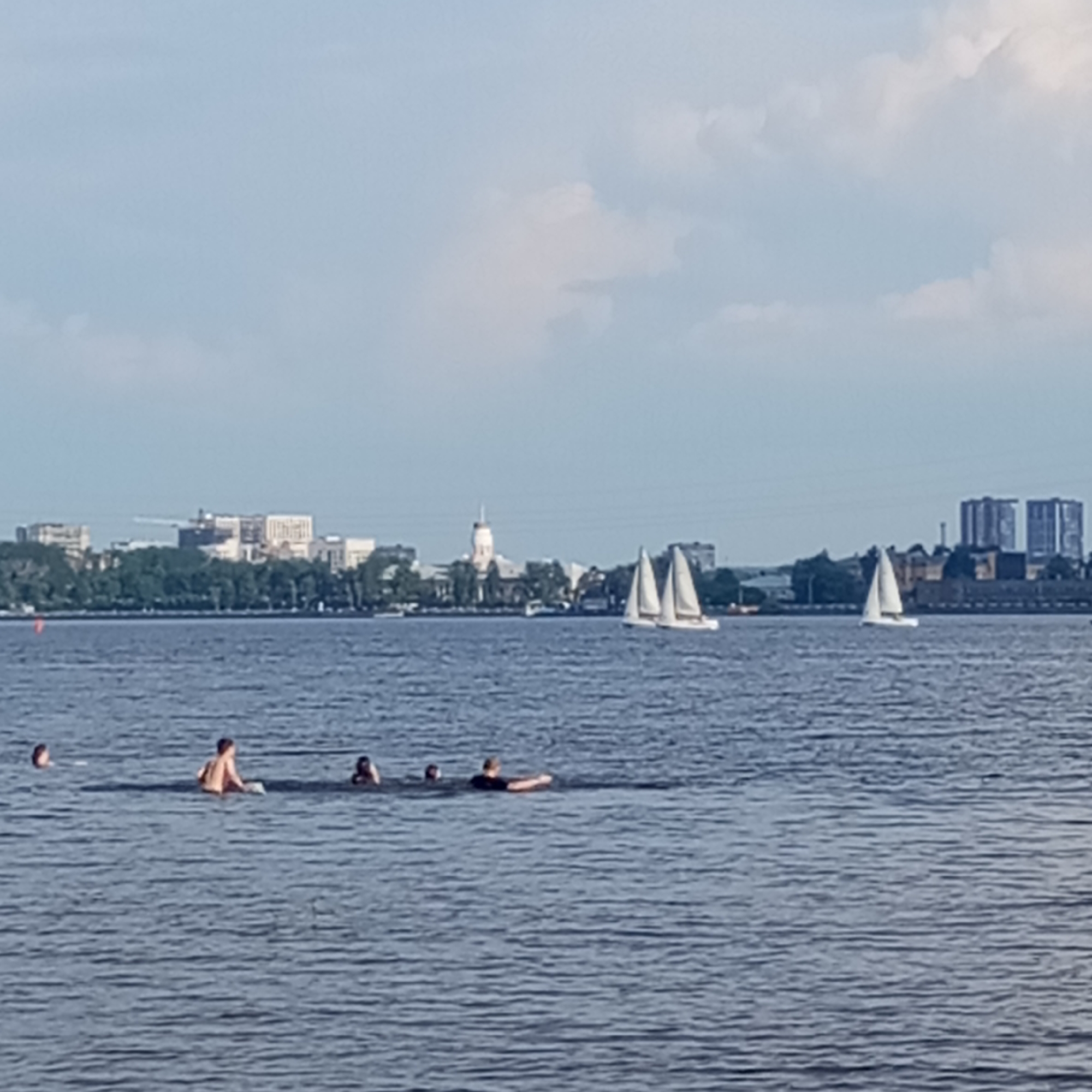 A wide shot shows a body of water, possibly a river or lake, with a city skyline in the background. Several people are swimming or wading in the water in the foreground. In the middle ground, there are several sailboats on the water. The city skyline features a variety of buildings, including some modern high-rises and older structures with domed roofs. Trees line the waterfront in front of the city. The sky is partly cloudy, with a hazy blue hue. The image appears to be taken on a bright day, possibly in the summer. It is not possible to determine the specific location within Izhevsk, Russia from the image alone, but the presence of a large body of water and the architectural style of the buildings suggest a possible waterfront area within or near the city.