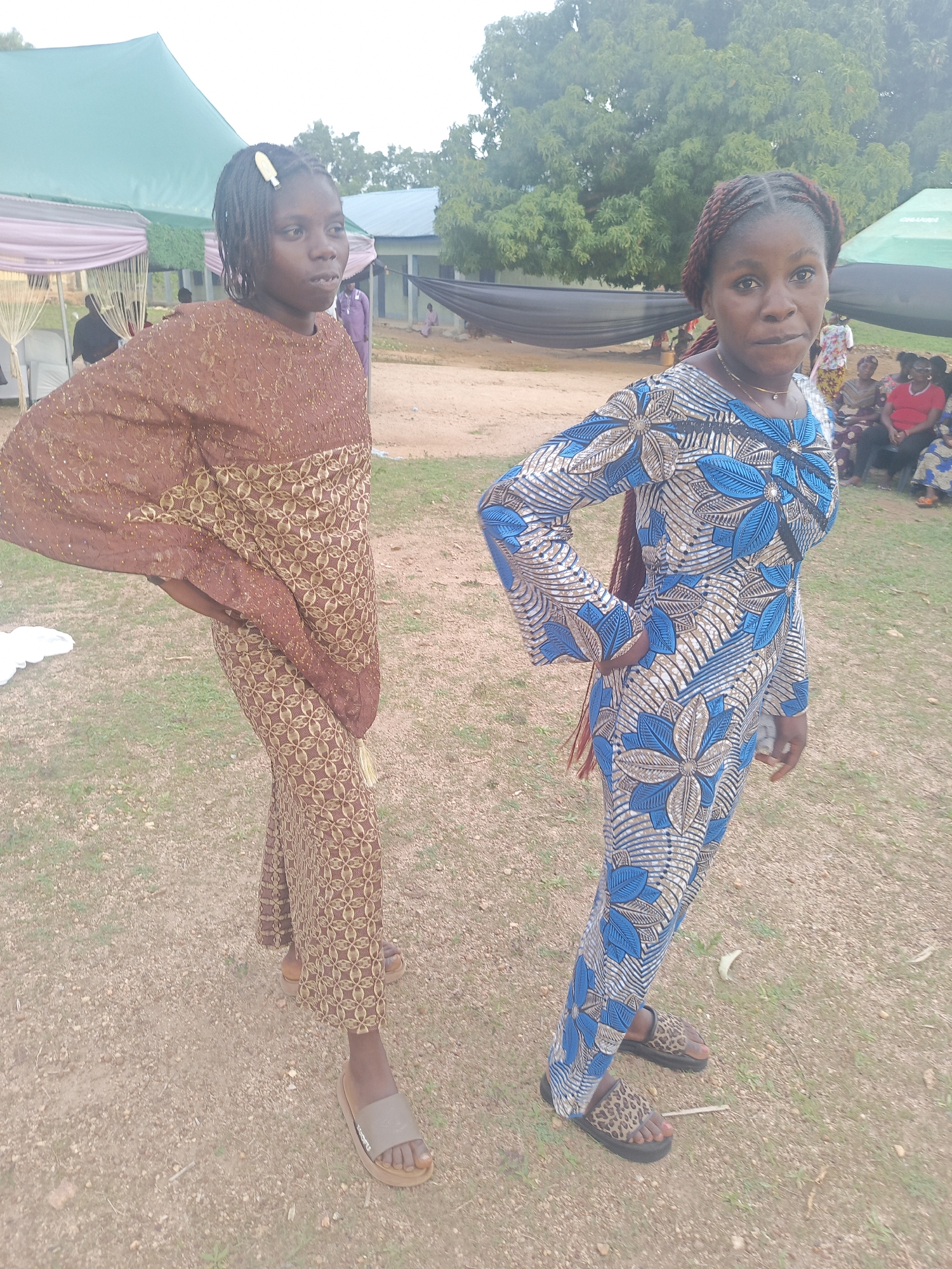 Two young women stand side-by-side in an outdoor setting in Unguwar Zaria, Nigeria. The ground is a mix of dirt and sparse grass. The woman on the left wears a brown, patterned dress and light-colored sandals. Her hair is braided and pulled back with a clip. The woman on the right wears a blue and white floral patterned dress and animal print sandals. Her hair is styled in red-tinted braids. Both women have one hand on their hips and are looking towards the camera. In the background, there is a green canopy tent, a building with blue trim, and a group of people seated. A large tree is visible behind the woman on the right.