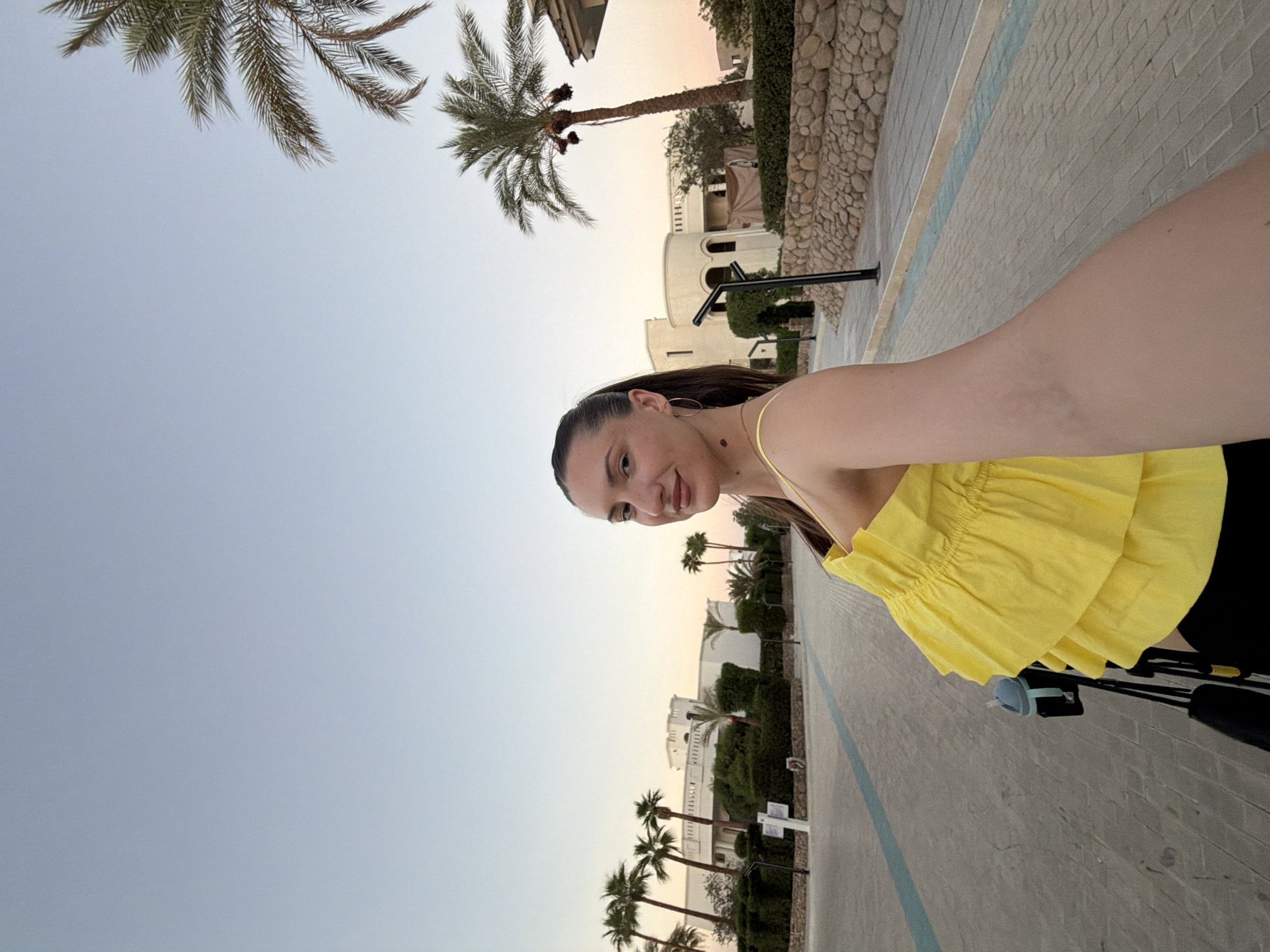 A woman is taking a selfie in an outdoor environment with palm trees and buildings in the background. She is wearing a bright yellow ruffled top and black pants. The background appears to be a resort or hotel complex, possibly in Sharm El Sheikh, Egypt, given the presence of palm trees and the architectural style of the buildings. The sky is a clear, pale blue, indicating it is likely late afternoon or early morning. She is also holding a stroller with a cup holder attached. The ground is paved with interlocking bricks.