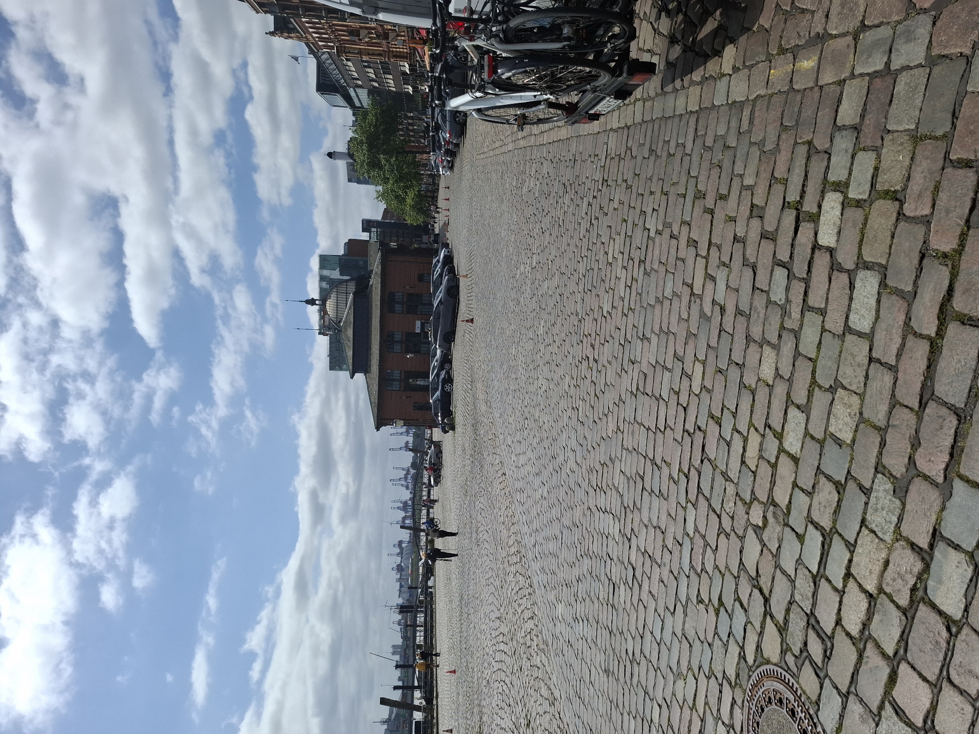 This is a daytime, eye-level, vertical, medium-wide shot of a cobblestone plaza in the Speicherstadt district of Hamburg, Germany, with a harbor and cranes visible in the background. The foreground is dominated by the textured, uneven cobblestone pavement, with a manhole cover on the left. Several cars are parked along the right side of the plaza. In the mid-ground, a brown brick building with a domed roof is prominent, and more parked cars are visible. Two individuals are walking across the plaza in the mid-ground, one holding an umbrella. Beyond the plaza, a body of water leads to a harbor filled with numerous cranes and industrial structures. The sky is a mix of blue and white clouds. On the far right, a trailer with two bicycles is attached to an unseen vehicle, partially visible. The scene depicts a public urban space with a mix of historic architecture and harbor infrastructure.