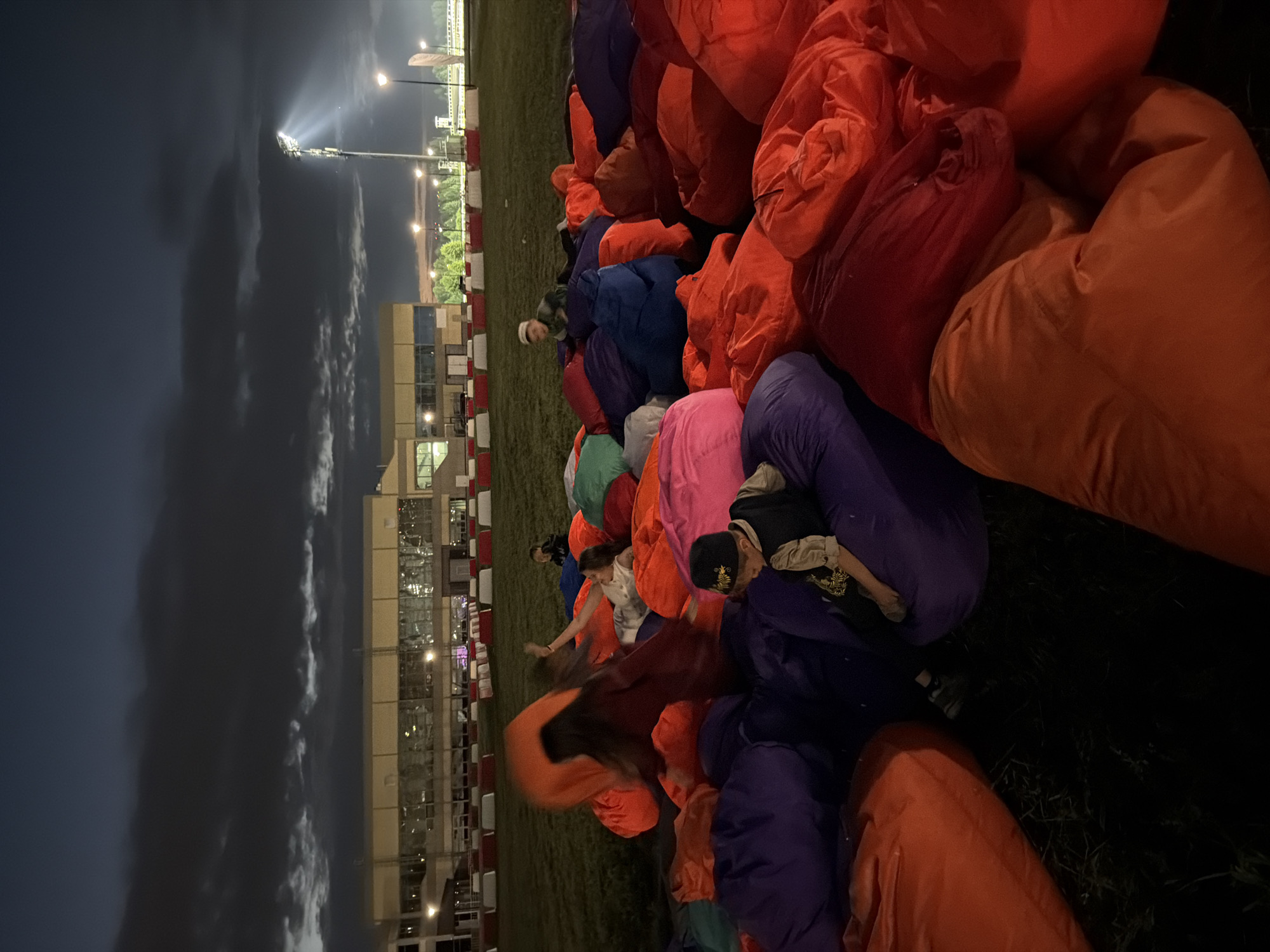 The image depicts a twilight scene on a grassy field with a building in the background. A large number of oversized, colorful beanbag chairs are scattered across the foreground and middle ground, in shades of orange, red, purple, pink, green, and blue. Several people are interacting with the beanbag chairs. In the middle ground, a person in an orange shirt is partially visible, appearing to be seated or interacting with a beanbag. To their right, another person is lying on a beanbag chair. In the foreground, a young boy wearing a vest and a cap is seated on a purple beanbag. Further back, another individual is visible sitting on a beanbag. The sky is dark with some clouds, and artificial lighting is present in the background, including a tall light post emitting a strong beam of light and streetlights. The building in the background appears to be a large, modern structure with many windows. There are also red and white barriers visible in the middle ground, possibly indicating a track or event area. The overall impression is of an outdoor gathering or event held in the evening.