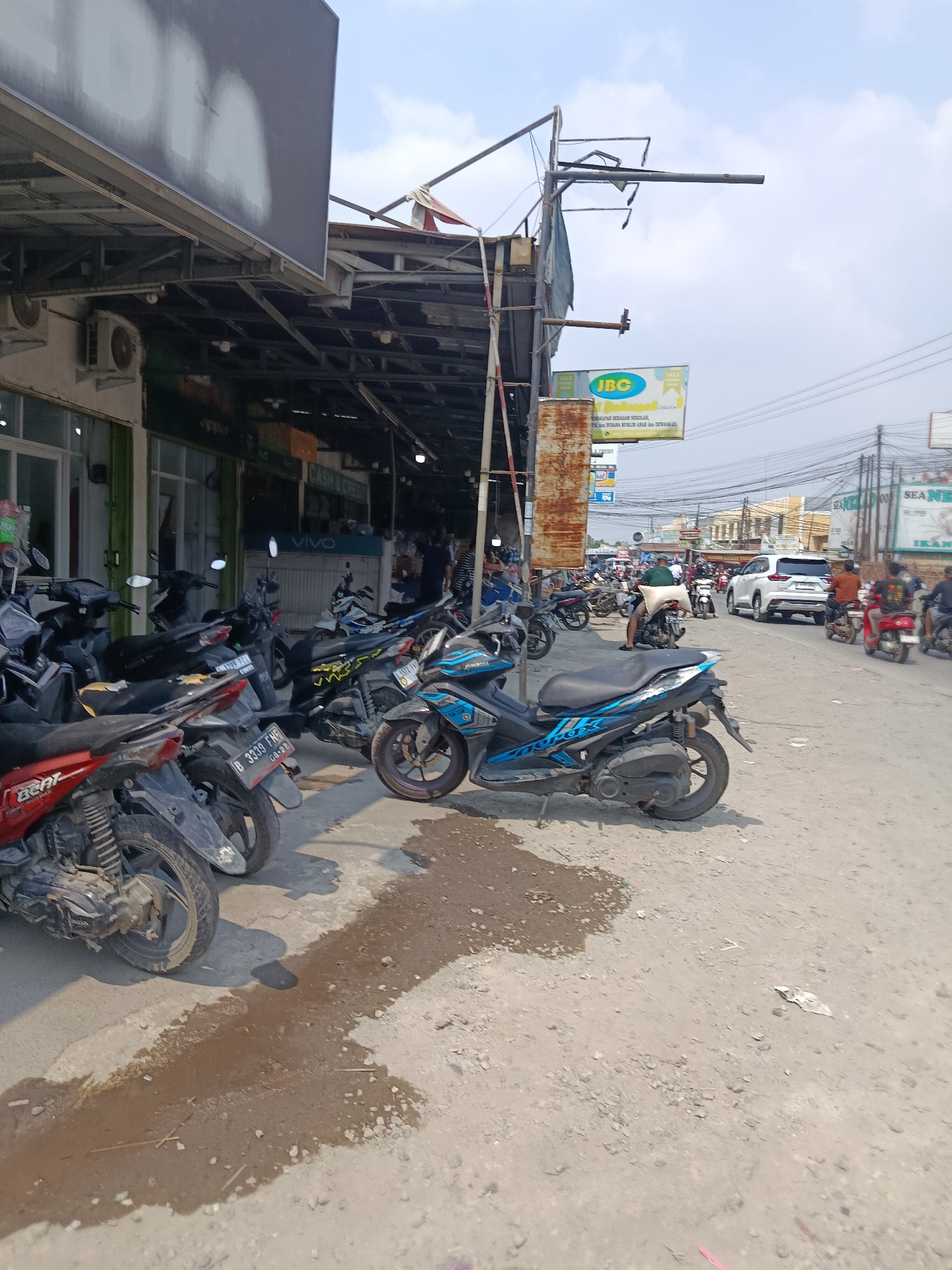 A street scene in Wanasari, Indonesia shows a row of parked motorcycles under a covered walkway. A white SUV and several other motorcycles are visible on the street, with a few people walking or riding. Buildings with awnings line the left side of the street, with signs for "JBC" and "VIVO" visible. The ground is a mix of dirt and gravel, with some wet patches. Overhead, a tangle of electrical wires crosses the sky, which is partly cloudy. The overall impression is a busy commercial area on a sunny day.