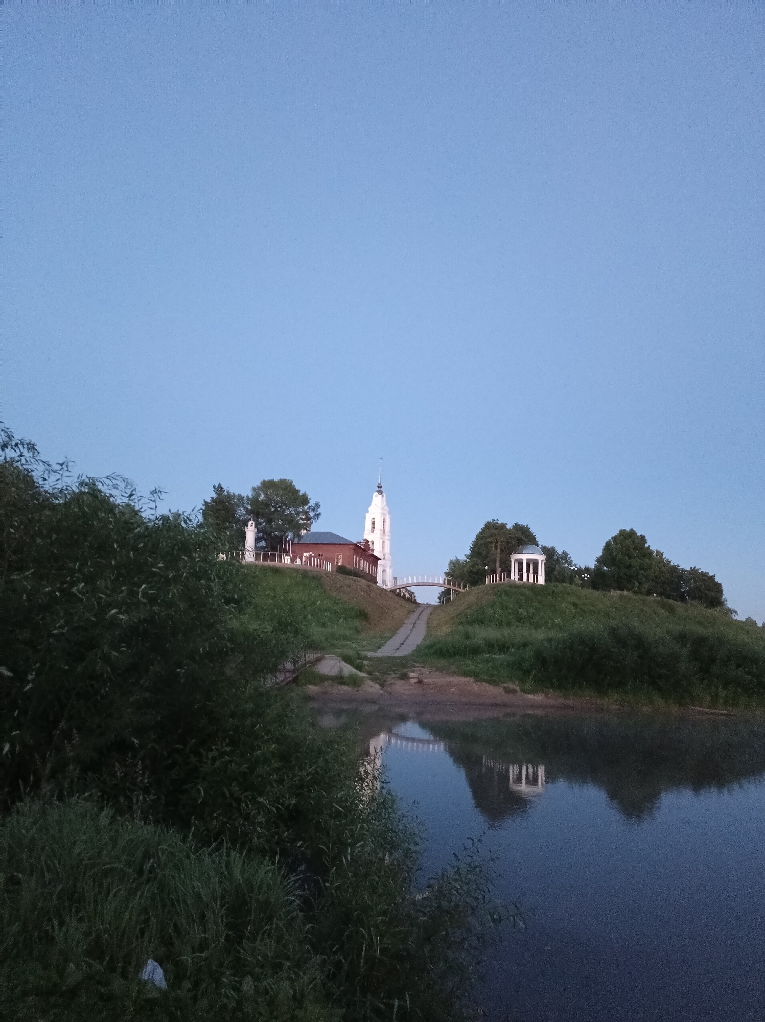 This is a photograph of a riverbank in Buy, Russia. In the background, on a hill, there is a white bell tower and a red brick building, likely a church, with a white spire. There is also a white rotunda structure on the hill, and a bridge connecting two parts of the hill. The sky is a clear, pale blue, indicating it is either early morning or late evening. The foreground is dominated by lush green foliage and grass. The river in the foreground reflects the sky and the structures on the hill. There are no people or animals visible in the image.