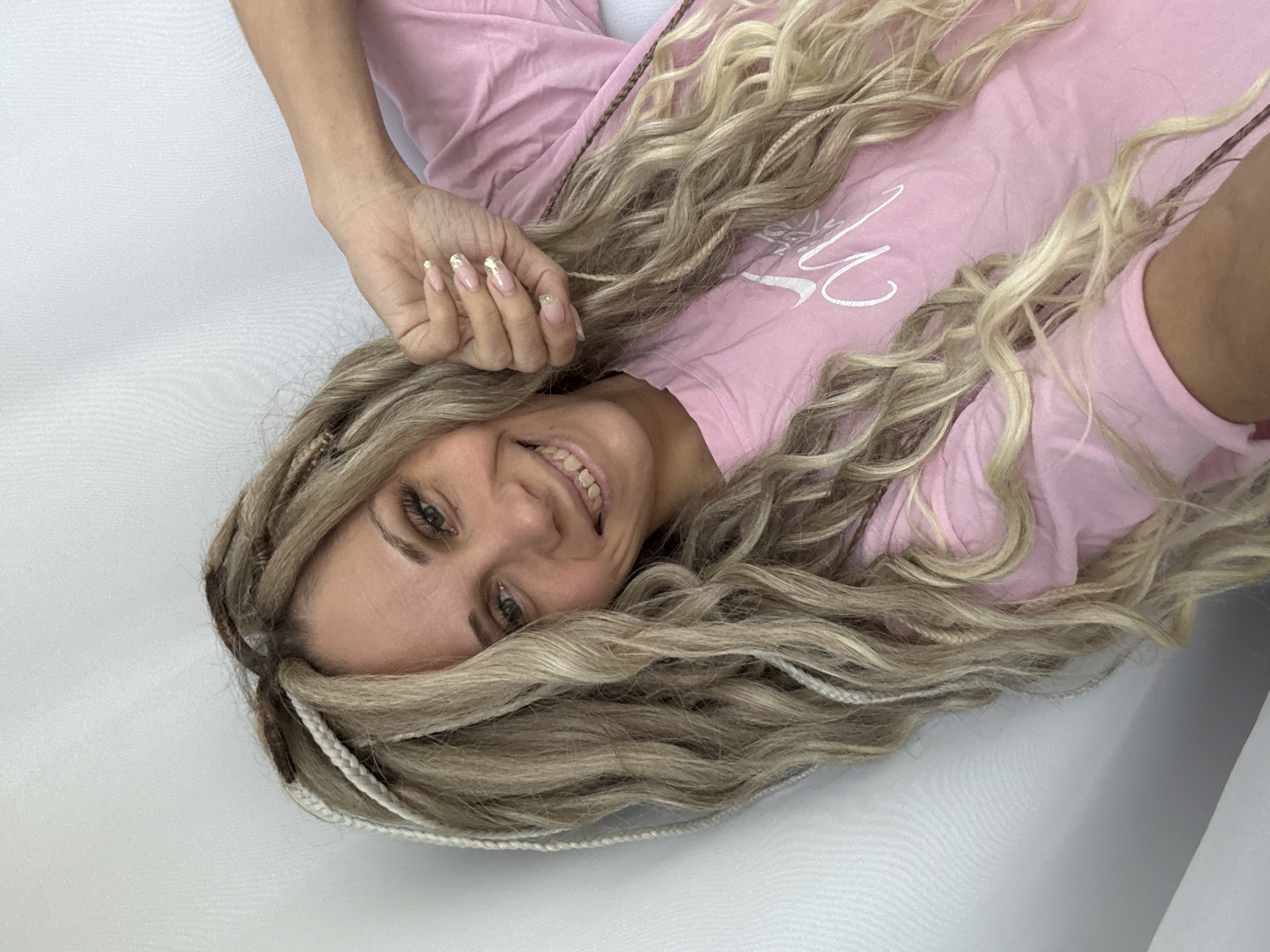 A woman is lying on a white surface. She has long, blonde, braided hair. She is wearing a pink shirt with a white logo. Her hand is near her face, and she has long fingernails painted with a white and gold design. She is smiling and looking at the camera. The location is Rusanovo, Russia.