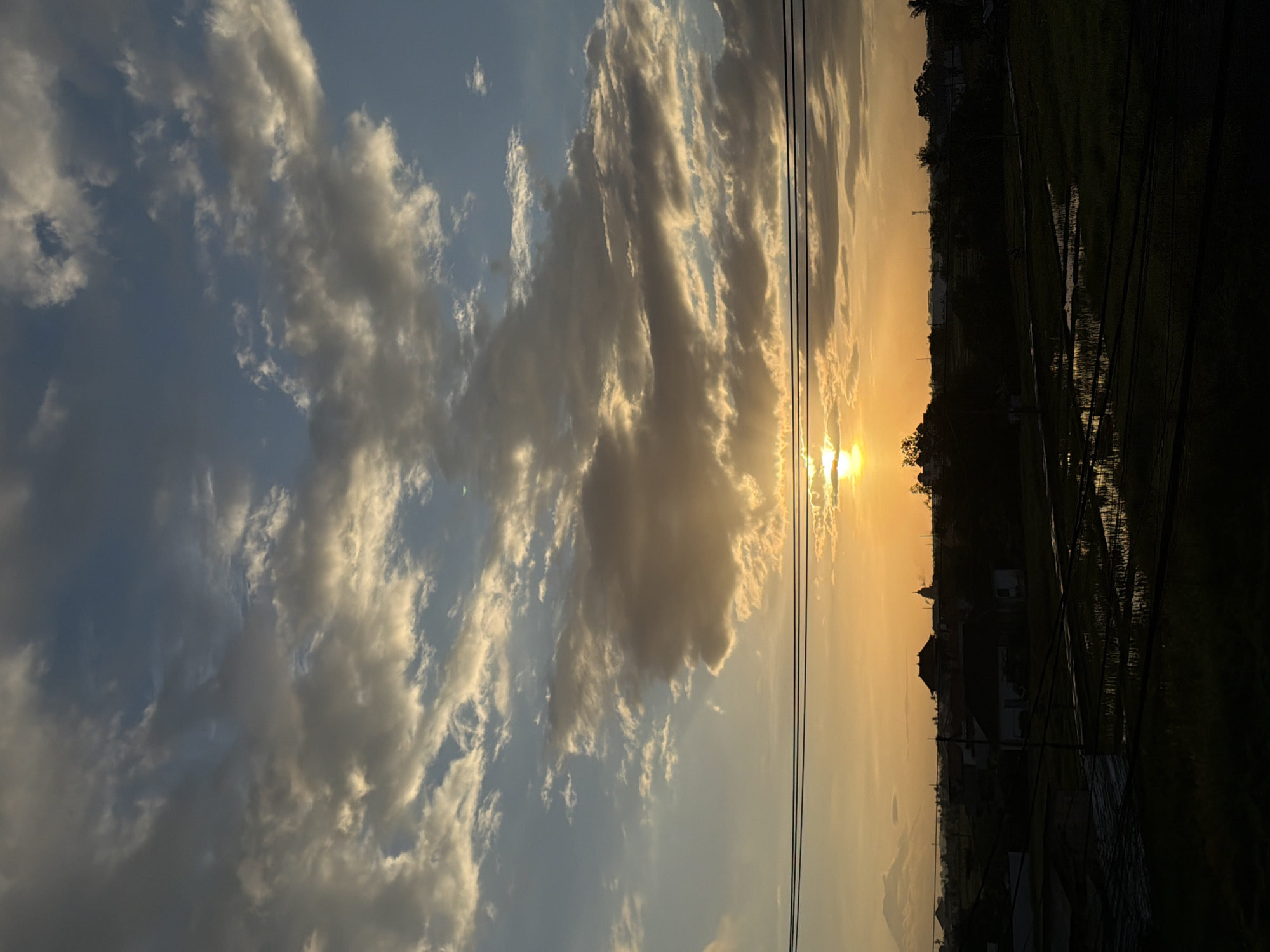 The image shows a landscape scene in Mengwi, Indonesia, captured during sunset. In the sky, there are patches of fluffy, light-colored clouds against a blue backdrop, with the sun partially obscured by a larger cloud, creating a golden halo effect. Utility lines run vertically across the view. Below the horizon, the foreground reveals a dark silhouette of trees and vegetation, possibly rice paddies, reflecting the orange-yellow light of the setting sun. There are a few buildings in the distance.