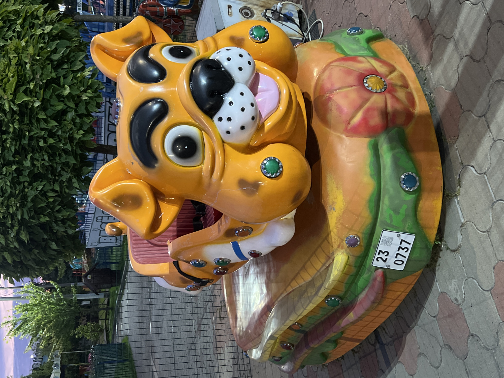 This is a medium shot of an orange amusement park ride shaped like a cartoon dog's head. The dog has large, black-rimmed eyes, a black nose with white polka dots, and a pink tongue sticking out. It's sitting on a base that resembles a hamburger, with layers of green, orange, and yellow. There are small, colored jewel-like accents around the base and on the dog's body. A license plate with the numbers "23 0737" is visible on the side of the base. The ride is situated on a paved walkway in what appears to be an amusement park or fairground. In the background, there are trees, a fence, and parts of other rides, suggesting an outdoor entertainment venue. The lighting indicates it might be dusk or evening. The specific location in Krasnodar, Russia, is not discernible from the image itself.