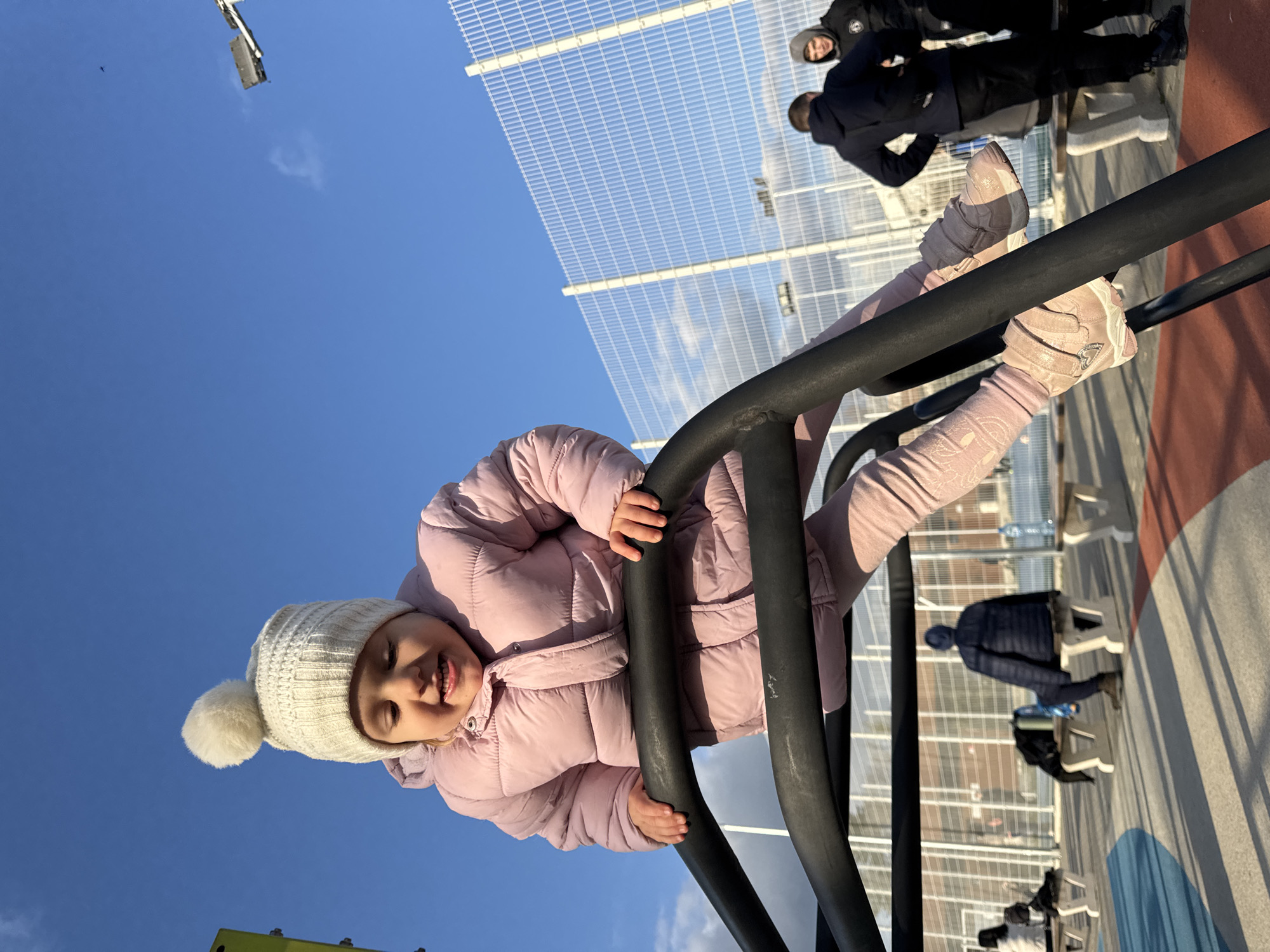 A girl in a pink puffer jacket and a white knit hat is hanging upside down on a black metal jungle gym. She is smiling. In the background, there is a light blue sky and a fence. People are standing and sitting in the background. The location is Varna, Bulgaria.