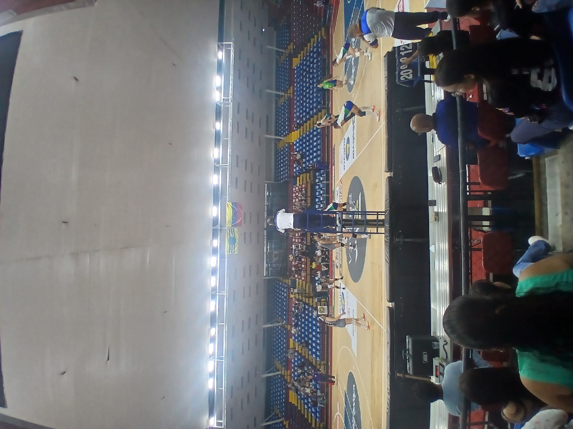 Here is a description of the image:
The image shows an indoor sporting event. It appears to be a volleyball game in progress within an indoor stadium. The stadium seating is blue and yellow and is partially filled with spectators. Spectators are visible along the side of the court. Players are seen on the court. There are scoreboards visible. The venue is illuminated by overhead lights. A flag is visible. The event location is Ciudad Guayana, Venezuela.