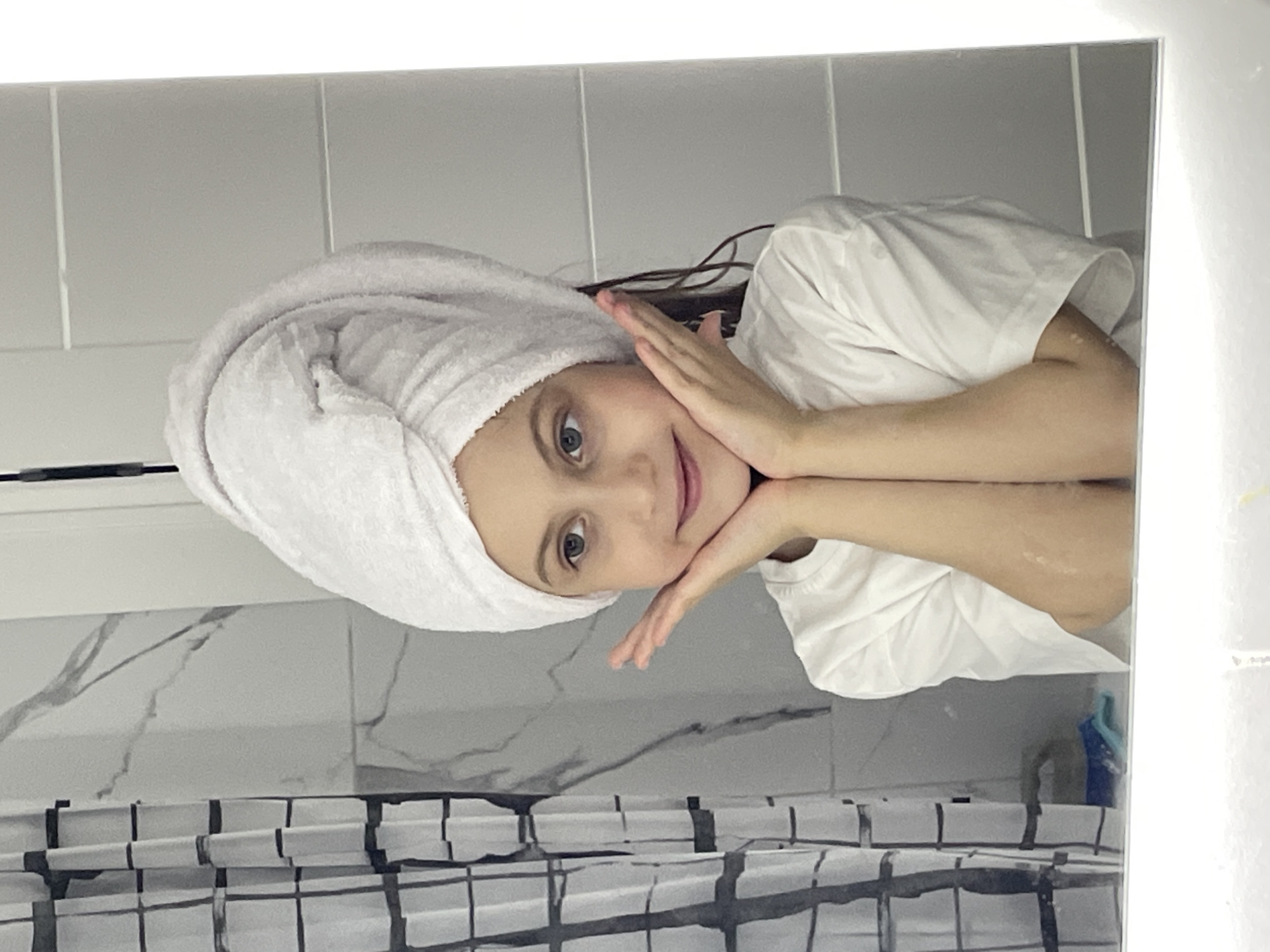 A young girl with fair skin, light brown hair, and grey eyes is reflected in a bathroom mirror. She is wearing a white t-shirt and a white towel wrapped around her head. Her hands are gently placed on either side of her face in a playful pose. The bathroom features light grey wall tiles and a black and white patterned shower curtain. The location in Izhevsk, Russia is not possible to determine from the given image.