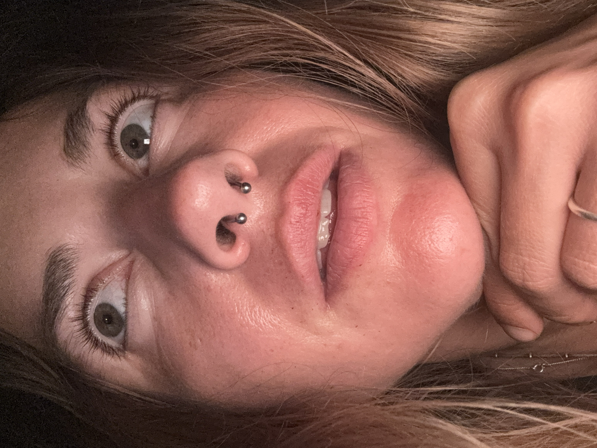 A close-up, low-angle indoor shot focuses on the face of a person, viewed from the nose down to the chin and chest. The subject has light skin with some freckles and reddish undertones. Their eyes are looking upwards and to the right, with long eyelashes. A septum piercing is visible on their nose. Their lips are slightly parted, revealing teeth. The subject's right hand is resting on their chin, with fingers slightly curled. A delicate silver necklace with small beads is visible on their chest. The background is dark, suggesting an indoor setting at night or in low light conditions.