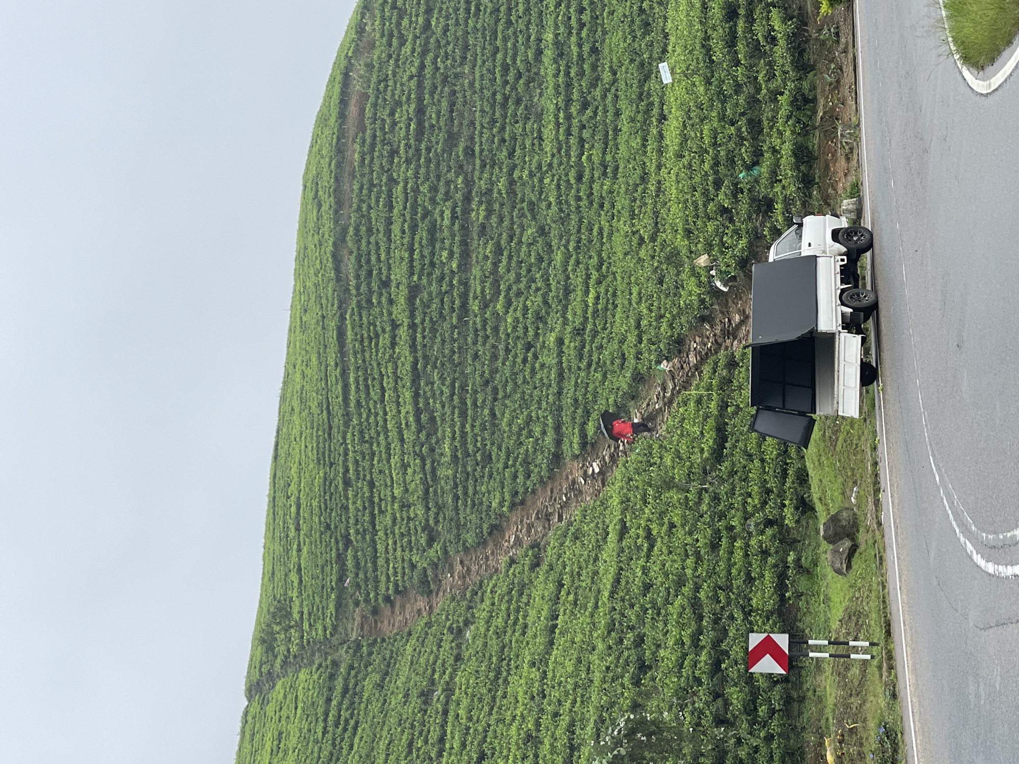 The image shows a lush, green hillside in Toppass, Sri Lanka, likely a tea plantation based on the dense rows of vegetation. A dirt path zigzags up the slope, and a person wearing a red top is walking along it. A white truck with a black cargo bed is parked on a paved road that runs alongside the bottom of the hill. A red and white road sign is visible near the road. The sky is overcast, suggesting a misty or rainy day.