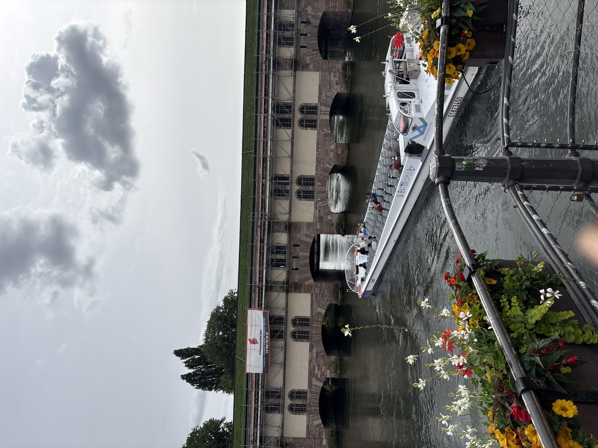 This is a vertical, eye-level, medium shot of the Barrage Vauban in Strasbourg, France, on an overcast day. A white tour boat named "BATORAMA" is moving down the Ill River, carrying passengers. The Barrage Vauban, a historic bridge with a series of arches, spans the river in the background. Scaffolding and a banner advertising "schoenberger" are visible on the structure, suggesting ongoing renovation work. In the foreground, a railing and a planter box filled with colorful flowers create a framing element. The sky is filled with dramatic, dark clouds.