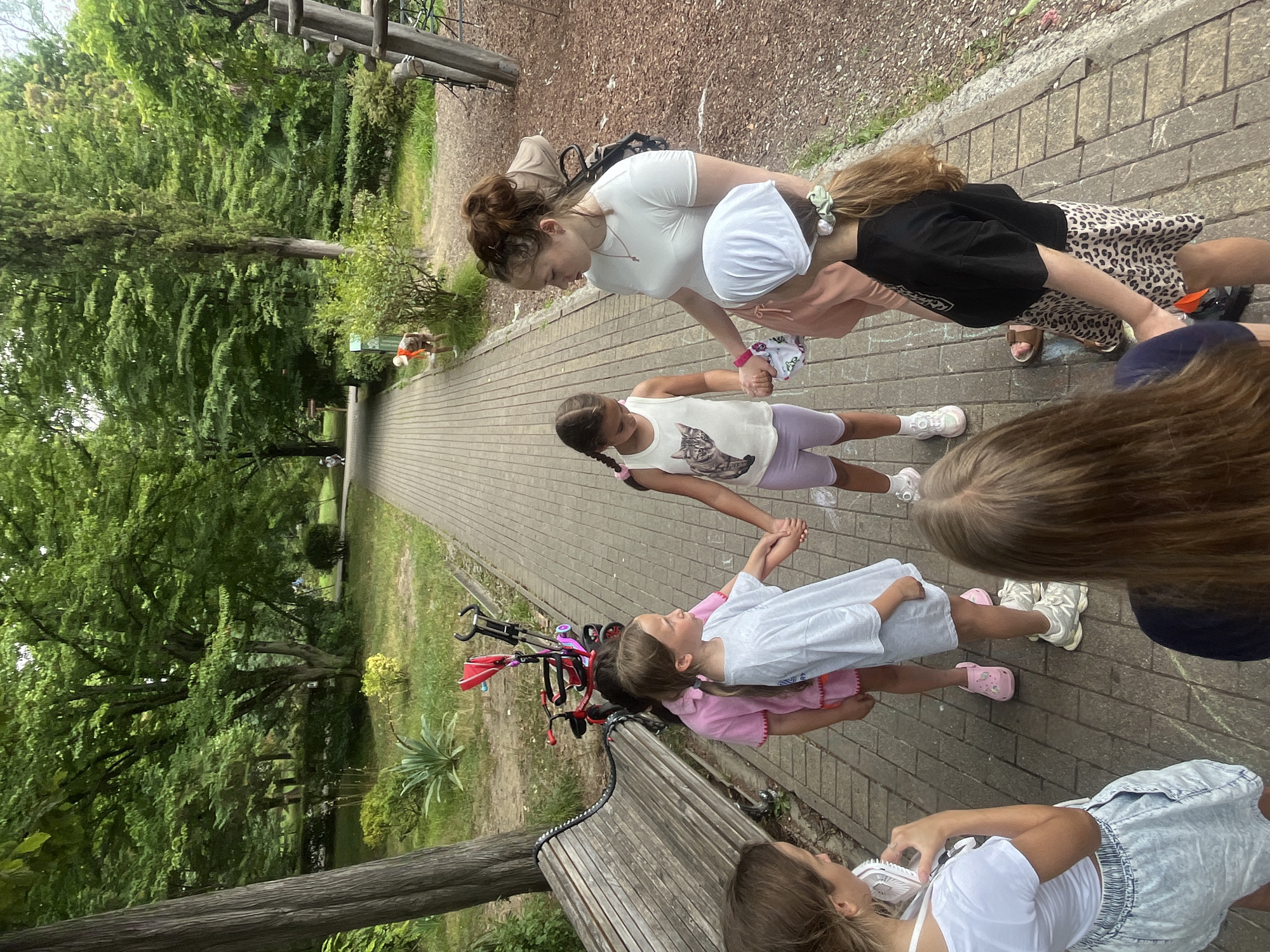 A group of children and one young adult are standing in a circle on a paved walkway in a park. The walkway is made of interlocking pavers and is bordered by grass and trees. In the background, there is a children's playground with wooden structures. A red stroller is parked near a wooden bench on the left side of the image. The young adult is wearing a white t-shirt and pink shorts. The children are wearing various casual outfits. The weather appears to be overcast. The location is likely a public park in Sochi, Russia, given the lush vegetation and playground equipment often found in such areas.