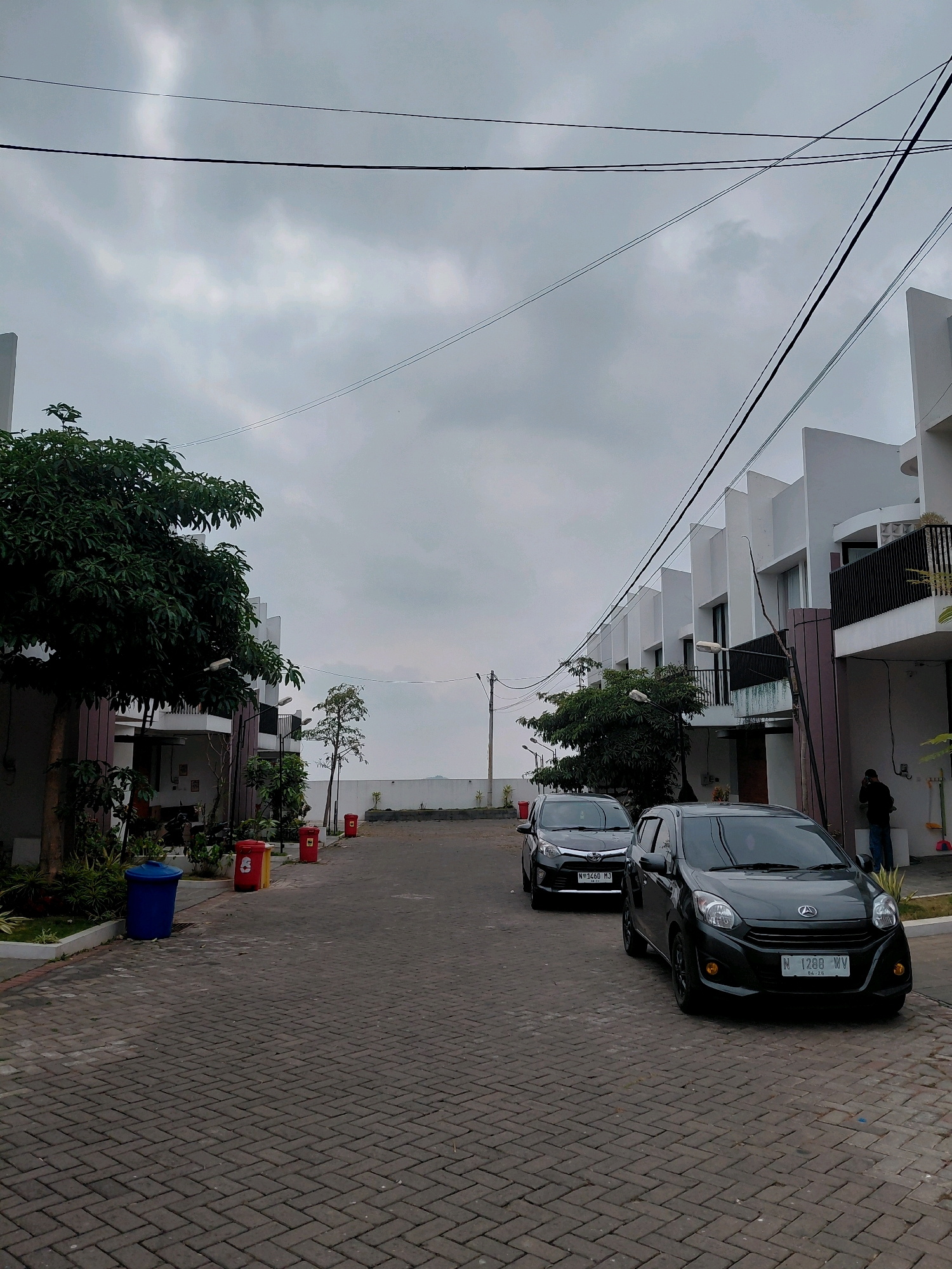 The image depicts a street scene in a residential complex in Batu, Indonesia. The overcast sky suggests a cloudy day. A row of modern, white houses with dark accents lines the right side of the street, featuring balconies and large windows. In the foreground, a paved driveway is made of interlocking dark grey bricks. Two dark-colored cars are parked on the driveway, one in front of the other. The car in the foreground is a dark grey Daihatsu with a license plate reading "N 1288 WV". The car behind it is a black Toyota with a license plate reading "N 3460 MJ".
On the left side of the street, there is a large, lush green tree, followed by more of the white houses. Several red trash bins and one blue trash bin are visible along the curb. In the distance, the street appears to continue towards a white wall, possibly marking the boundary of the complex. A person in dark clothing is standing near the entrance of one of the houses on the right, seemingly engaged in a phone call. Utility wires are strung overhead, crossing the frame from left to right. The overall impression is that of a quiet, suburban residential area.