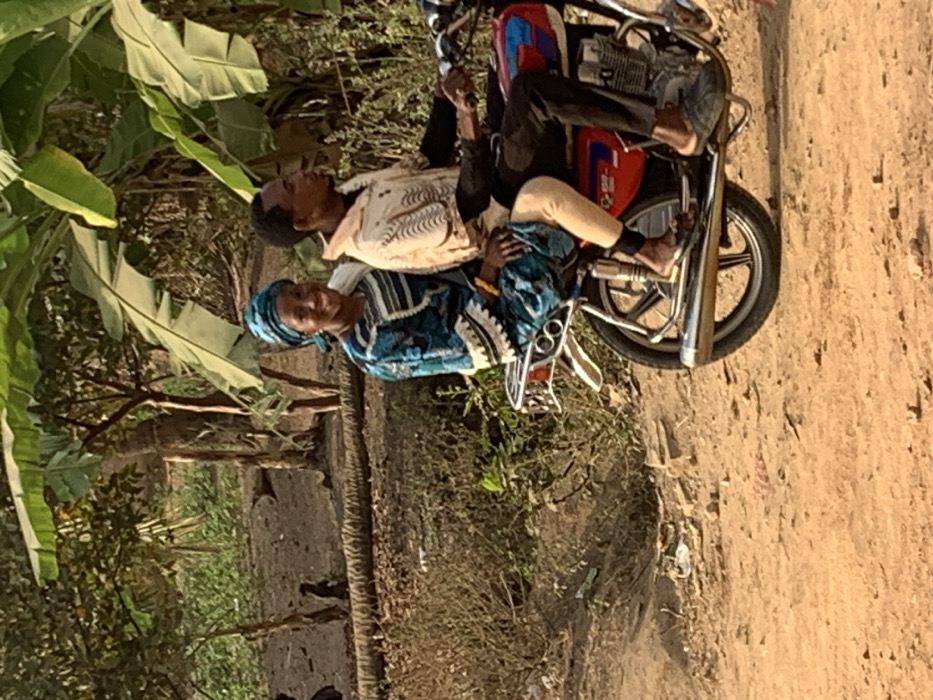 A man and woman sit on a parked motorcycle on a dirt road. The man wears a light-colored shirt and dark pants, while the woman is dressed in a blue and white patterned garment and headwrap. The motorcycle has a red gas tank and chrome accents. Lush green foliage is visible on the left side of the image, with dry brush and dirt ground in the middle.