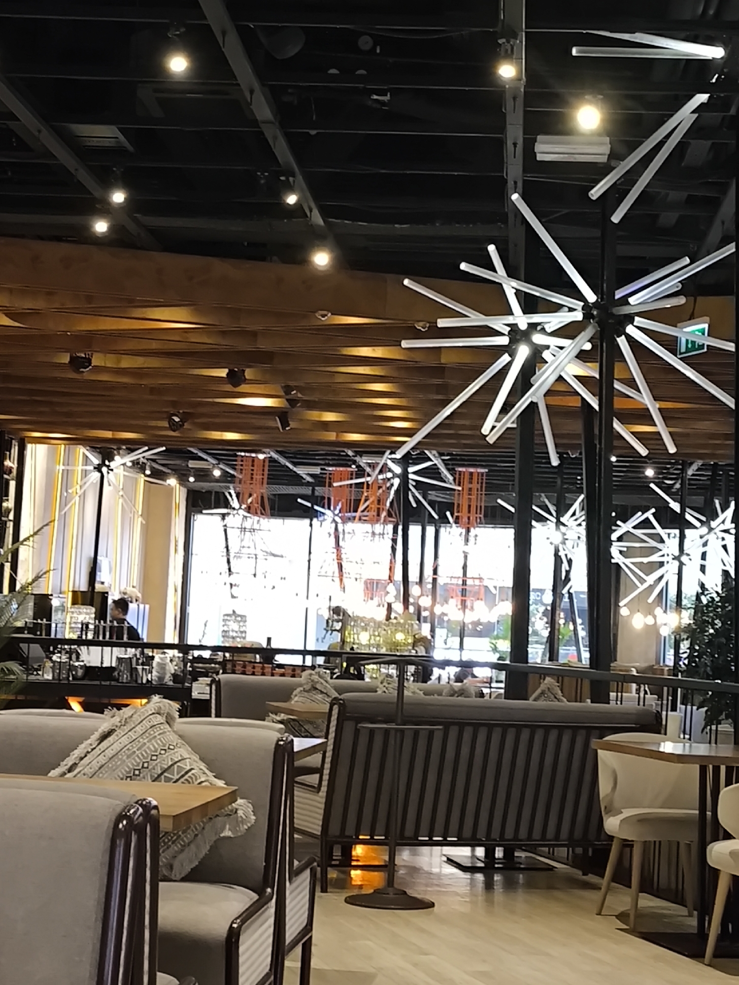 The image depicts the interior of a cafe located in Khimki, Russia. The cafe appears to be modern and spacious, with a high ceiling featuring exposed black beams and a wooden structure. Several pendant light fixtures with radiating white lights are prominently displayed. The cafe is furnished with comfortable seating arrangements, including sofas with decorative pillows and tables with chairs. A railing separates the seating area from a higher level, where a person can be seen working behind a counter. Large windows provide ample natural light and offer a view of the outside. The overall atmosphere is relaxed and contemporary.