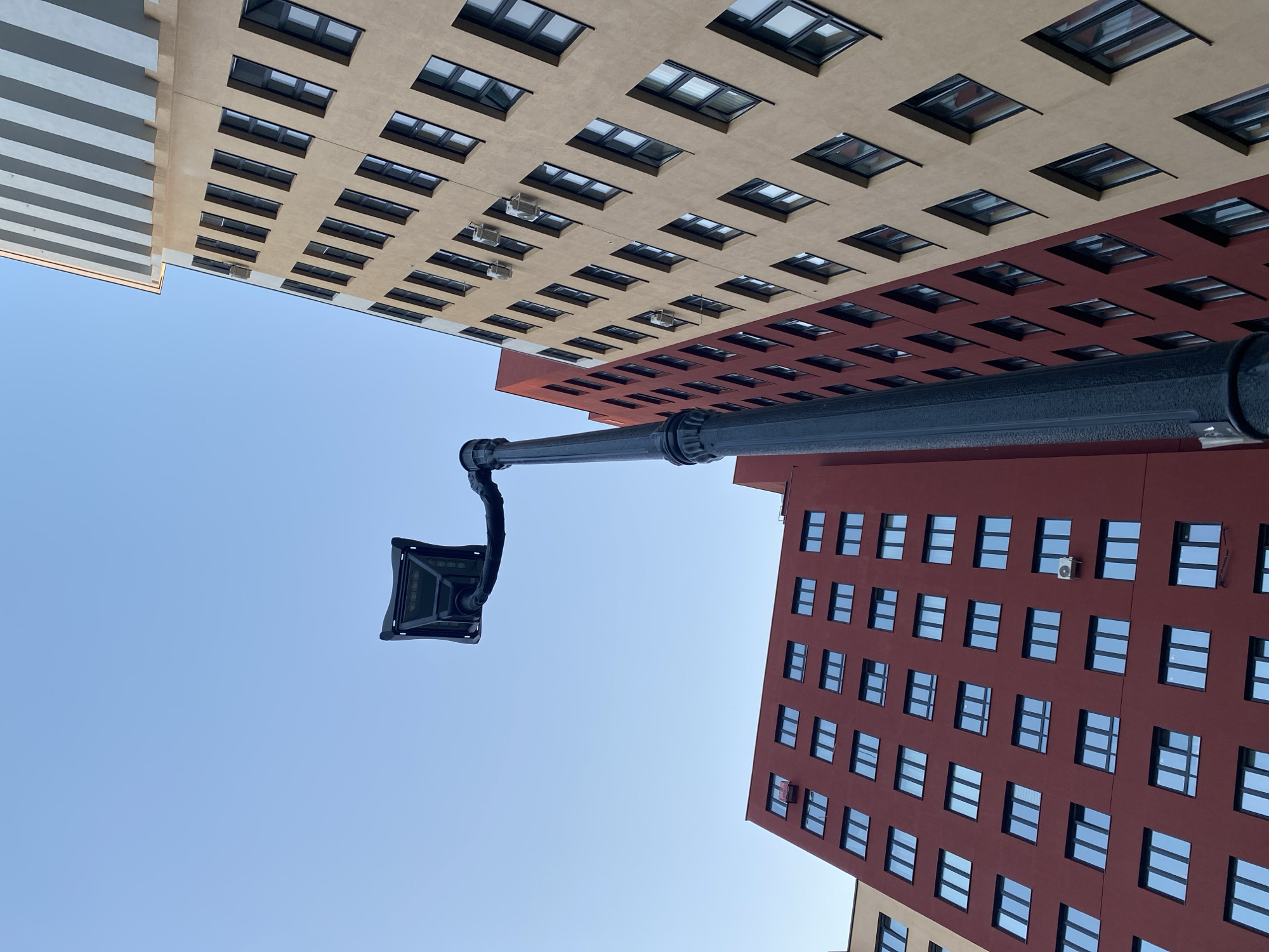 The image shows a view looking up at buildings in Yekaterinburg, Russia. A black lamppost with a square fixture is prominently positioned in the foreground. Two buildings flank the lamppost: one with a light beige facade and another with a red facade, both adorned with rows of windows. The sky is clear and blue.