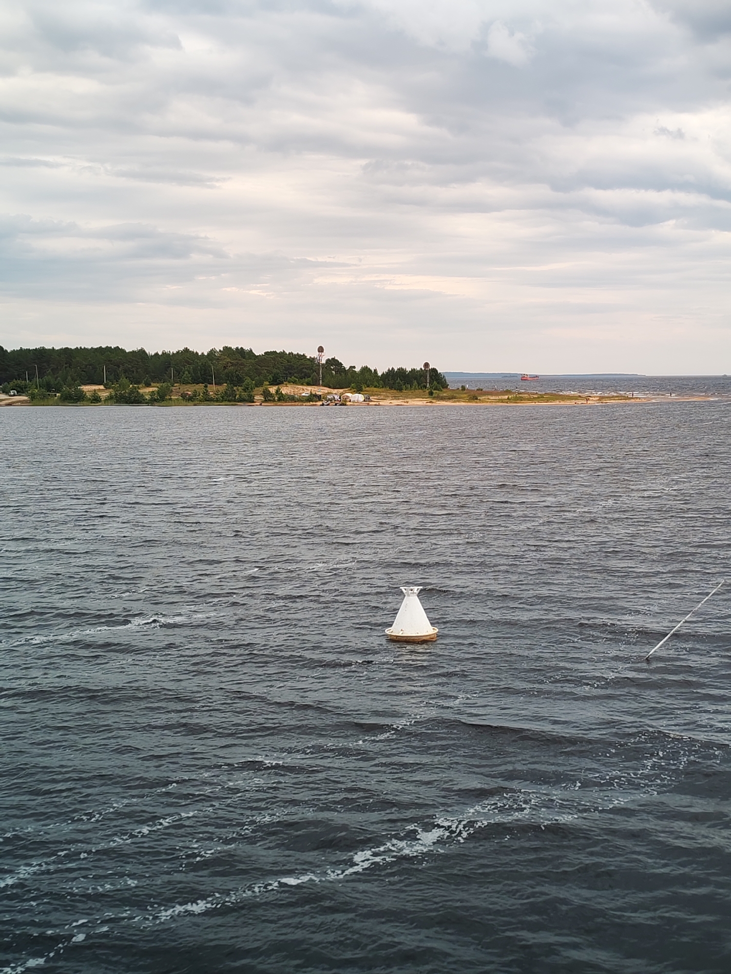 The image depicts a wide, dark body of water with a choppy surface, suggesting a breezy day. In the foreground, a white conical buoy with a yellow base floats on the water. A white pole extends diagonally from the right side of the frame into the water. The middle ground features a sandy shoreline lined with a dense forest of green trees. On the shoreline, there are a few structures, including what appears to be a lighthouse or tower and some smaller buildings. In the distance, a large red ship is visible on the horizon. The sky is overcast with gray clouds. The scene appears to be a view from the water looking towards the shore. The description of Shalsky, Russia, is not explicitly represented in the visual elements of the image.
