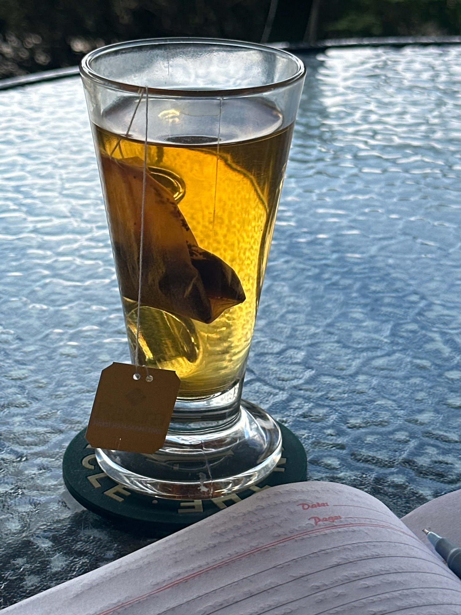 A close-up, eye-level shot shows a glass of amber-colored tea with a tea bag steeping inside, resting on a dark green coaster with embossed text, possibly "Kageshwori Manohara, Nepal." The glass is on a textured glass tabletop that reflects the ambient light, creating a rippled pattern across the surface. In the foreground, a portion of a notebook with lined pages and red ruling is visible, along with a pen. The background is out of focus, showing dark foliage. The scene suggests a moment of quiet contemplation or work outdoors.