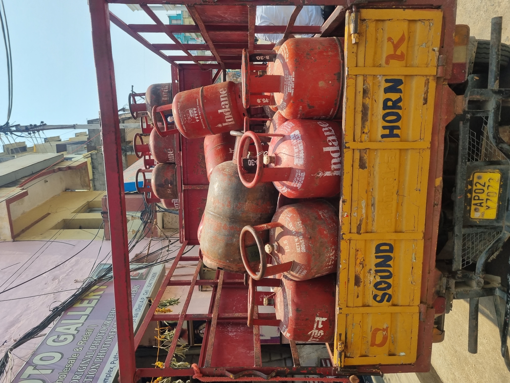 The image displays the rear view of a weathered, yellow and red open-bed delivery truck, situated in Puttaparthi, Anantapur District, Andhra Pradesh, India. The truck is heavily laden with approximately 15 to 20 red Liquefied Petroleum Gas (LPG) cylinders, predominantly of the "Indane" brand. These cylinders exhibit visible wear, including dirt, rust, and scuff marks, with some labels indicating a "NET WT. 14.2 KG" and referencing the "Pradhan Mantri Ujjwala Yojana."
The truck's yellow tailgate is stenciled with the words "SOUND HORN" in dark blue capital letters, along with a red "K" symbol and an orange circular symbol. Its license plate, visible beneath the tailgate, reads "AP02 Y7072". The red upper frame of the truck is also weathered, and some dried garlands are affixed to its left side.
In the background, a multi-story building with a light pink facade is visible, featuring a partially discernible sign that reads "PHOTO GALLERY" and "Puttaparthi Complex," confirming the location. Numerous black electrical and communication cables crisscross the scene overhead, against a clear blue sky. A partial view of an individual wearing a white shirt is visible through the truck's red side railing on the right. The ground beneath the truck is an unpaved, light-colored surface.