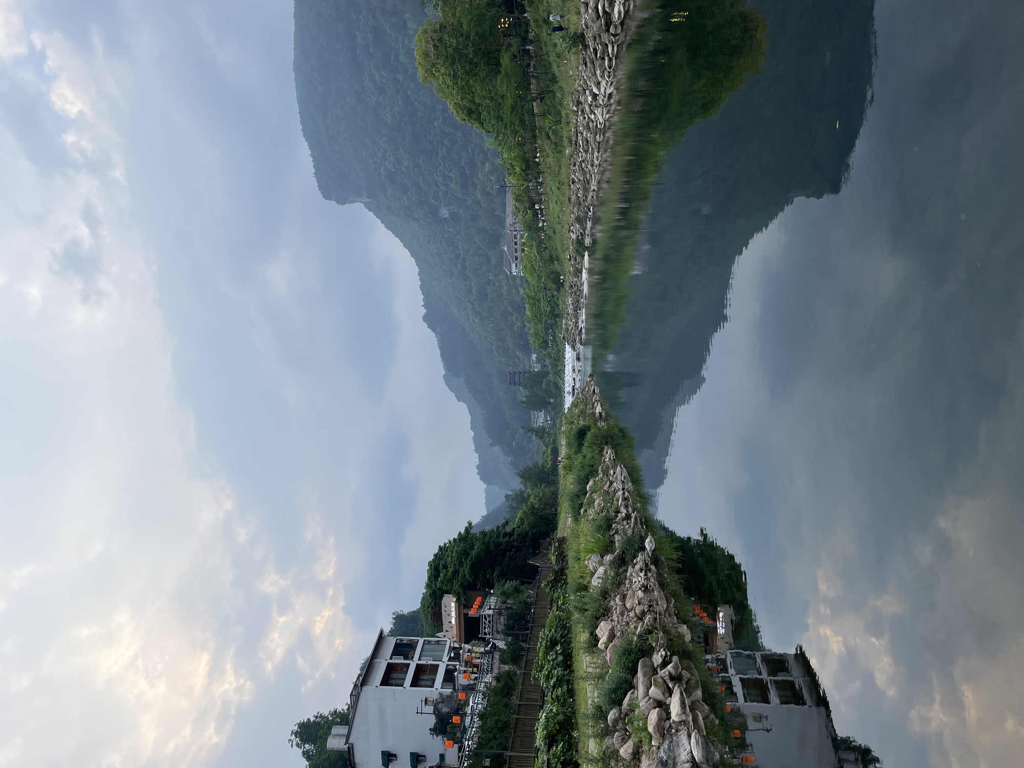 The image shows a landscape scene, likely a reflection in water, given the inverted symmetry. The scene features a mountain range in the background under a cloudy sky, with the clouds reflected in the water. A building with windows and what appears to be decorations or lights is visible. A rock-lined shore separates the land from the water. The location is suggested to be Wulingyuan, China.