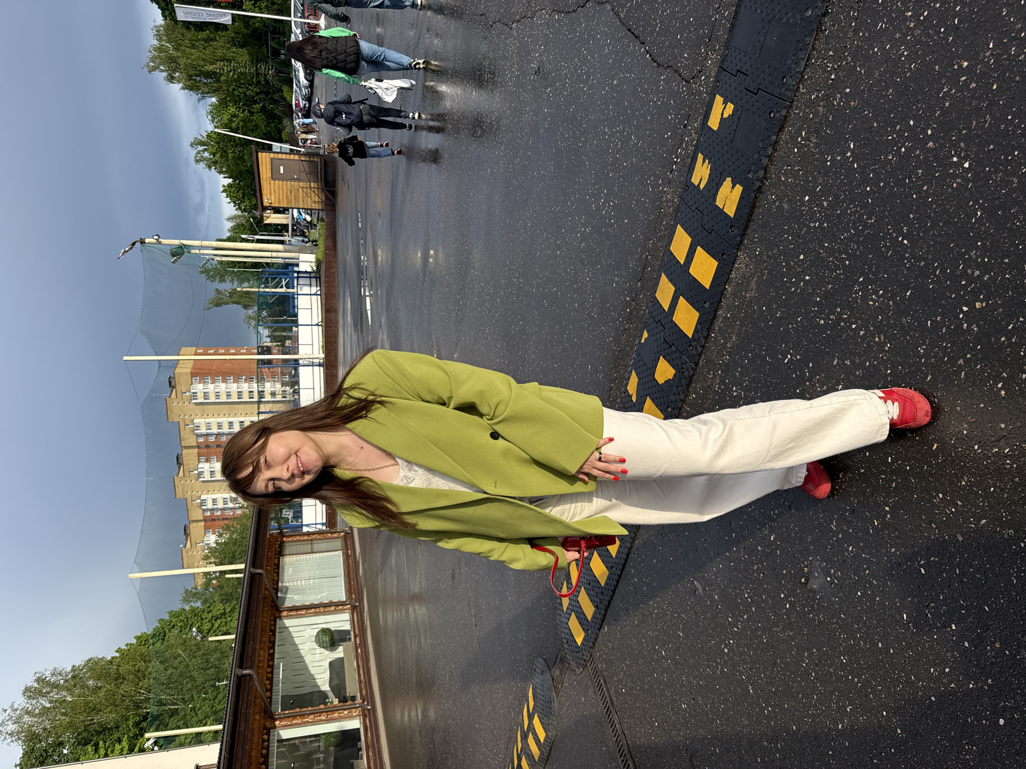 A young woman stands on a wet, asphalt-paved area with black and yellow speed bumps, possibly a parking lot or street. She wears a light green blazer over a white top, white pants, and red sneakers. She is holding a small red bag. In the background are buildings, including a multi-story apartment building and a single-story structure with windows, as well as trees, netting, and other people walking in the distance. The sky is overcast. The location is Peresvet, Russia.
