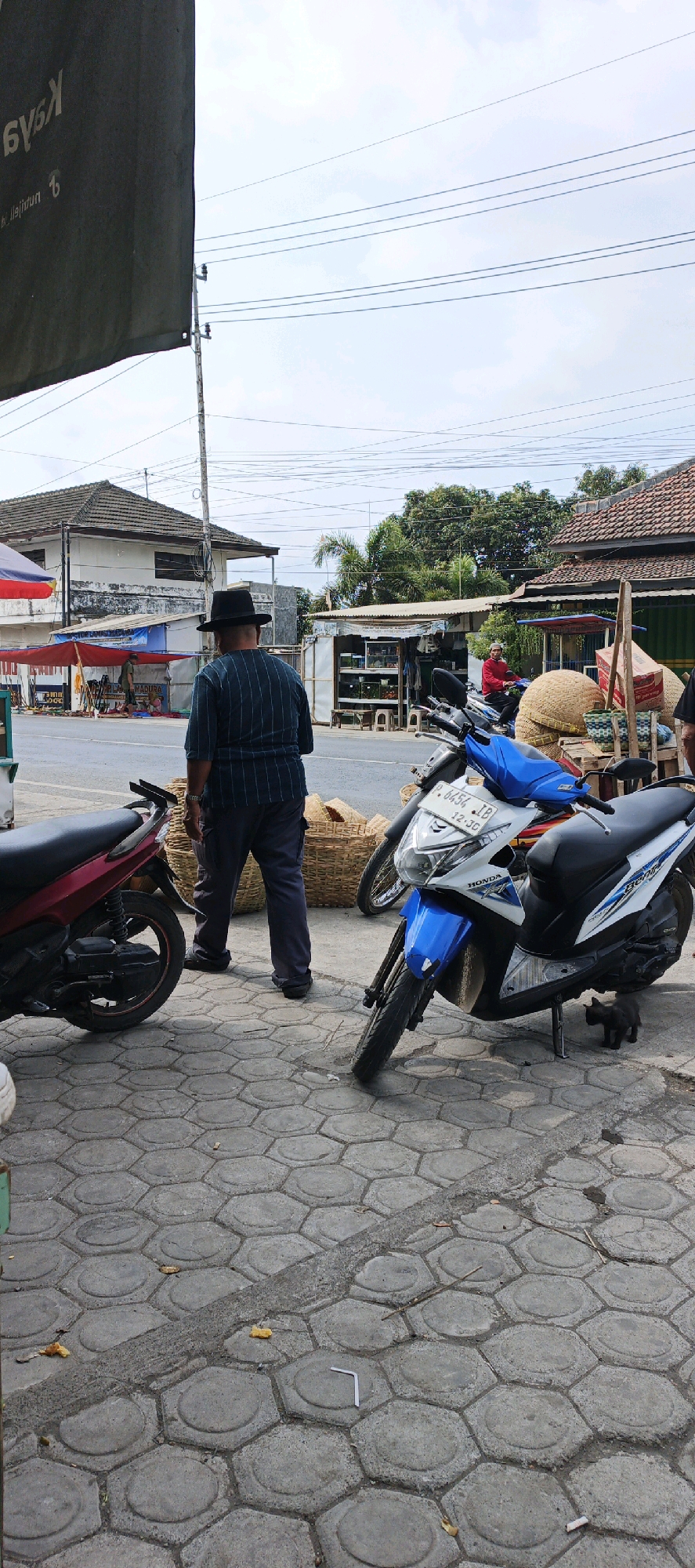 In the city of Kasiyan, Indonesia, on a paved sidewalk with hexagonal concrete blocks, a man wearing a black hat and a vertically striped blue and dark blue shirt stands with his back to the camera. He is facing a street with several shops and stalls on the opposite side. To his right are two motorcycles parked on the sidewalk. The closest is a white and blue Honda motorcycle, and behind it is a red motorcycle. A small black cat is visible on the sidewalk near the front wheel of the white and blue motorcycle. In the background, a man in a red shirt rides a motorcycle down the street. Several large woven baskets are stacked behind the motorcycles. Power lines are visible in the sky overhead.