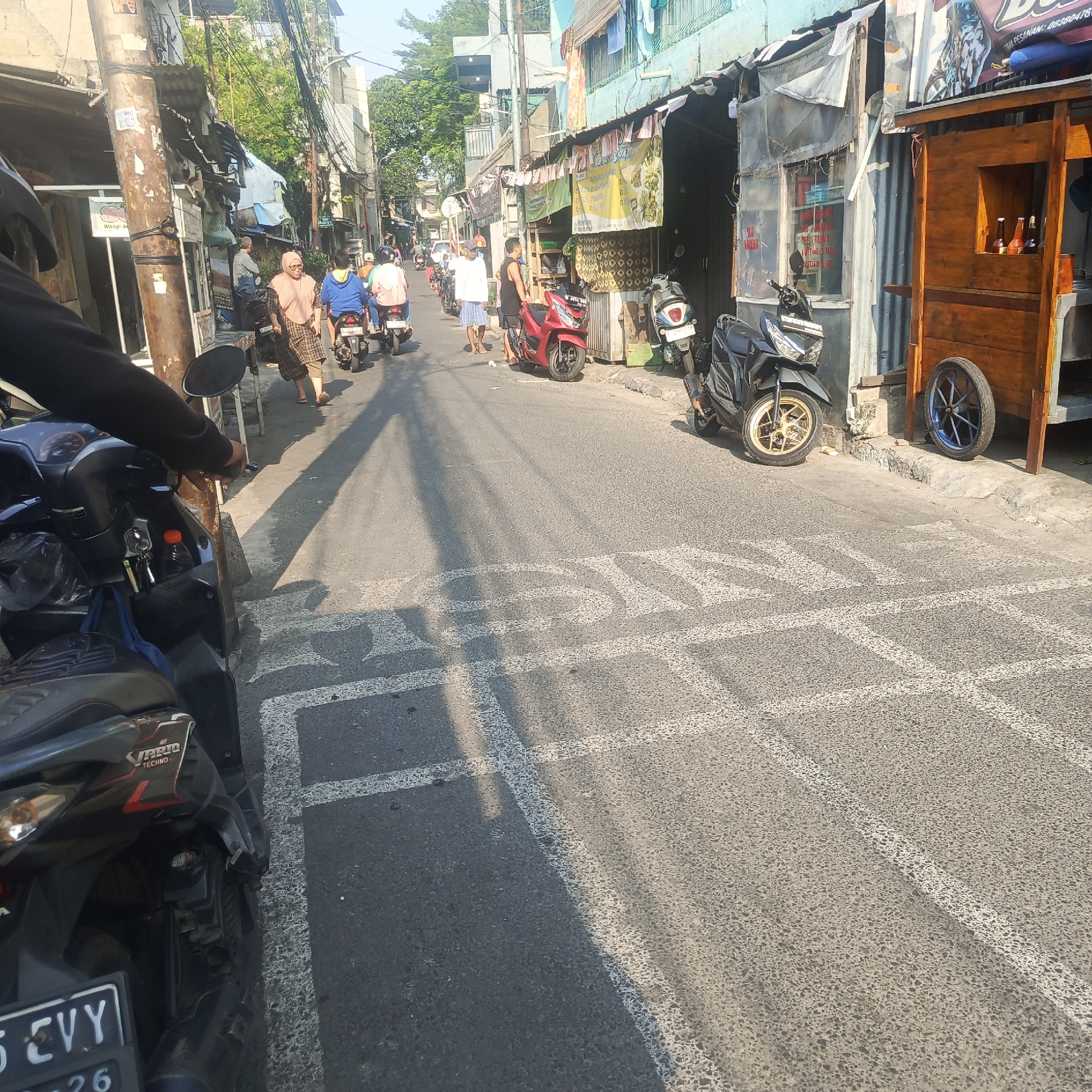 The image depicts a street scene in South Jakarta, Indonesia, likely during daylight hours as indicated by the strong sunlight and shadows. The street is narrow and lined with buildings that appear to be a mix of shops and residences, some with makeshift awnings and signage. The architecture suggests an urban, possibly somewhat informal, commercial or residential area.
Several motorcycles are present, indicating their common use as transportation in this region. One motorcycle is in the foreground, partially obscuring the view of the person operating it, who is wearing dark clothing. Other motorcycles are seen further down the street, some carrying riders. People are walking and standing on the sidewalks and in the street, suggesting a moderate level of pedestrian activity. One woman in the middle ground is wearing a headscarf and a patterned dress.
The street surface is asphalt and shows some wear. There are painted white lines on the road, possibly indicating pedestrian crossings or parking zones. A wooden cart with wheels, perhaps for selling goods, is parked on the side of the street, with bottles visible inside. The overall impression is of a bustling, everyday street in a densely populated urban environment.
