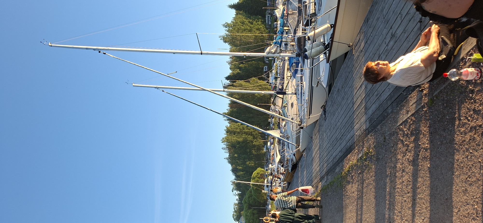 In Salpakangas, Finland, a marina scene unfolds under a clear blue sky. A wooden boardwalk runs along the edge of the water, where several sailboats are docked. Two individuals, one in a green jumpsuit and the other in a striped shirt, walk along the boardwalk. The person in the striped shirt carries a white plastic bag. Further down the boardwalk, a woman with short, reddish-brown hair sits on a bench, bathed in sunlight, with a bottle of pink liquid and a juice box on the ground beside her. Behind the boats, a dense line of trees forms the backdrop, with some parked cars and what appear to be RVs visible in the distance. The overall atmosphere suggests a warm, late afternoon.