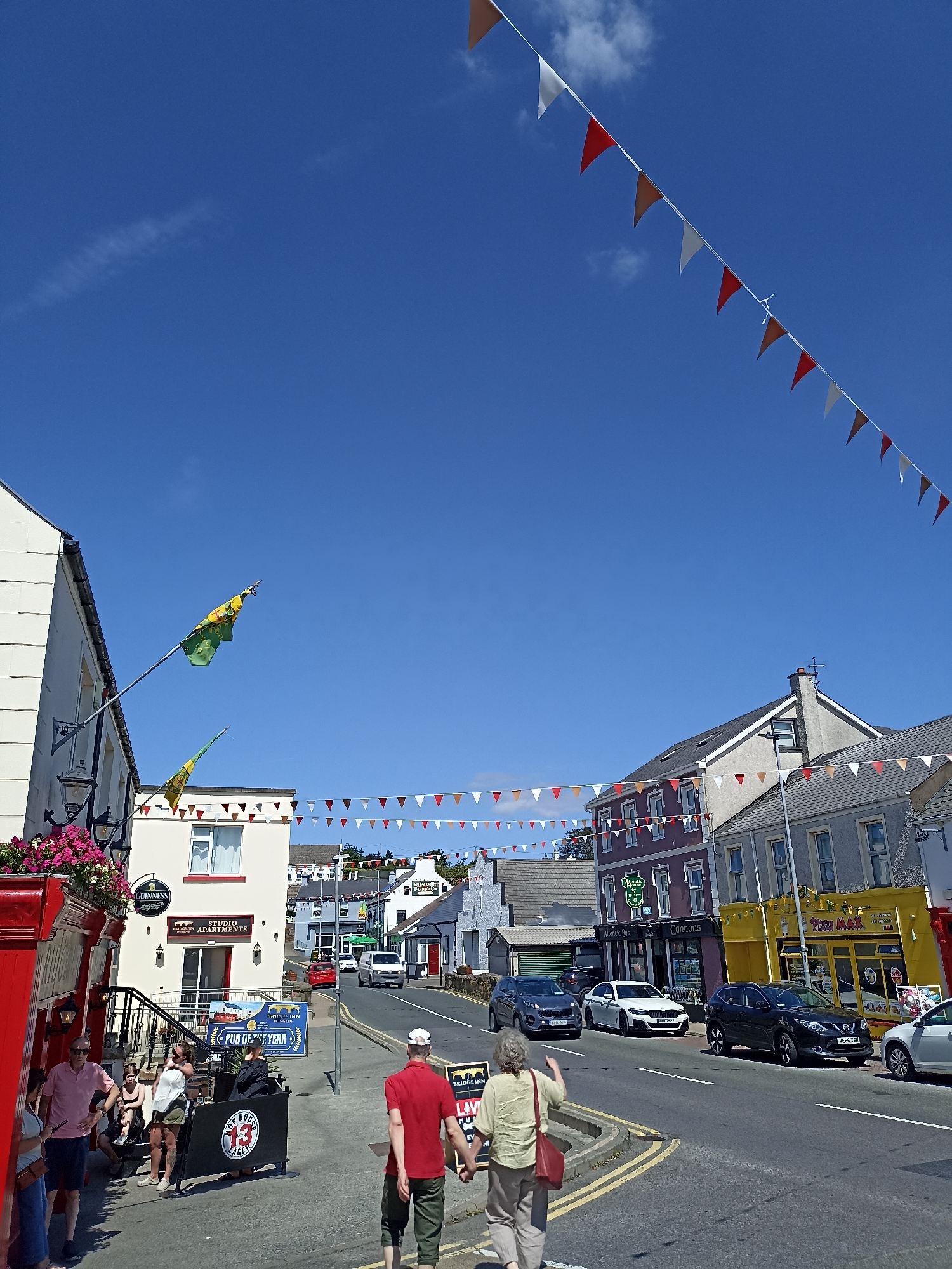 The image captures a sunny day on a street in Dungloe, Ireland, likely during a festive occasion given the abundant triangular bunting strung across the street. The street is lined with buildings of various colors and architectural styles. On the left, a red building with a Guinness sign and flowers above the entrance stands prominently. Adjacent to it, a cream-colored building is labeled "STUDIO APARTMENTS."
Several people are visible on the sidewalk. A group is gathered near the red building, some standing and conversing, while others are seated. A couple in the foreground walks away from the camera, with the man in a red shirt and the woman in a light green top and light-colored pants carrying a red bag.
The street itself has a clear blue sky above, with a few wispy clouds. Cars are parked along the street and some are in motion, including a white van, a white sedan, and a black SUV. Businesses with visible signage such as "Minsters Bar," "Connors," and "Pizza Max" are interspersed along the street. Two flags, green with yellow emblems, are mounted on poles extending from one of the buildings. The overall impression is a lively town center on a bright day.