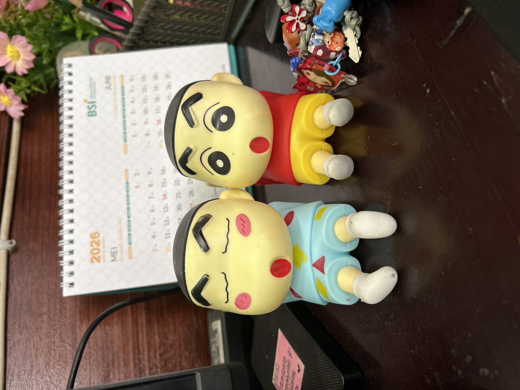 The image shows a desk with several items. Two cartoon figurines, one in a red shirt and yellow shorts, and the other in a light blue outfit with pink cheeks, lie next to each other. A small desk calendar showing the months of May and June 2026 sits nearby. Also visible is a basket with various items, a flower arrangement, and a collection of keychains. A black cord and the corner of a monitor are visible at the bottom of the frame. A pink sticky note is attached to a device at the bottom. The surface is a dark brown wood. The text "DIVISI mangamat lan" is printed on the sticky note, which indicates the location is likely Lhokseumawe, Indonesia.