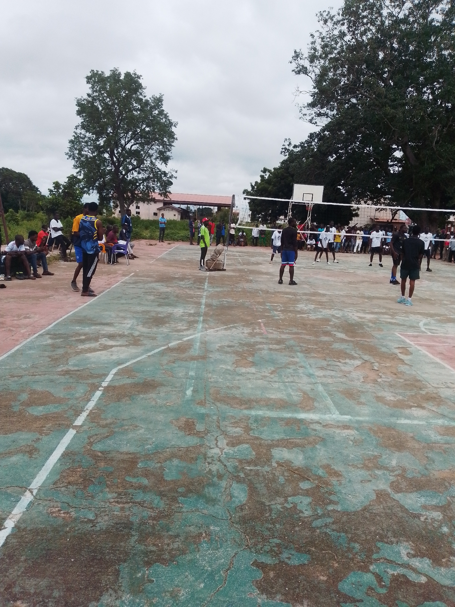 This is a medium shot, outdoor, eye-level photograph taken in Wukari, Nigeria, depicting a group of people on a weathered, outdoor volleyball court. The court surface is primarily a teal-green color with areas of dirt and wear, marked with white lines. In the background, a volleyball net is stretched across the court, and a basketball hoop with a backboard is visible.
Several individuals are present. On the court, some are actively engaged in a volleyball game, with players in various positions on either side of the net. Other individuals are standing around the court, observing the game or socializing. Some are seated on a bench or chairs on the side of the court.
The environment appears to be somewhat rural or semi-urban, with trees and a building visible in the background. The sky is overcast, suggesting a cloudy day. The overall scene conveys a sense of community gathering and recreational activity.