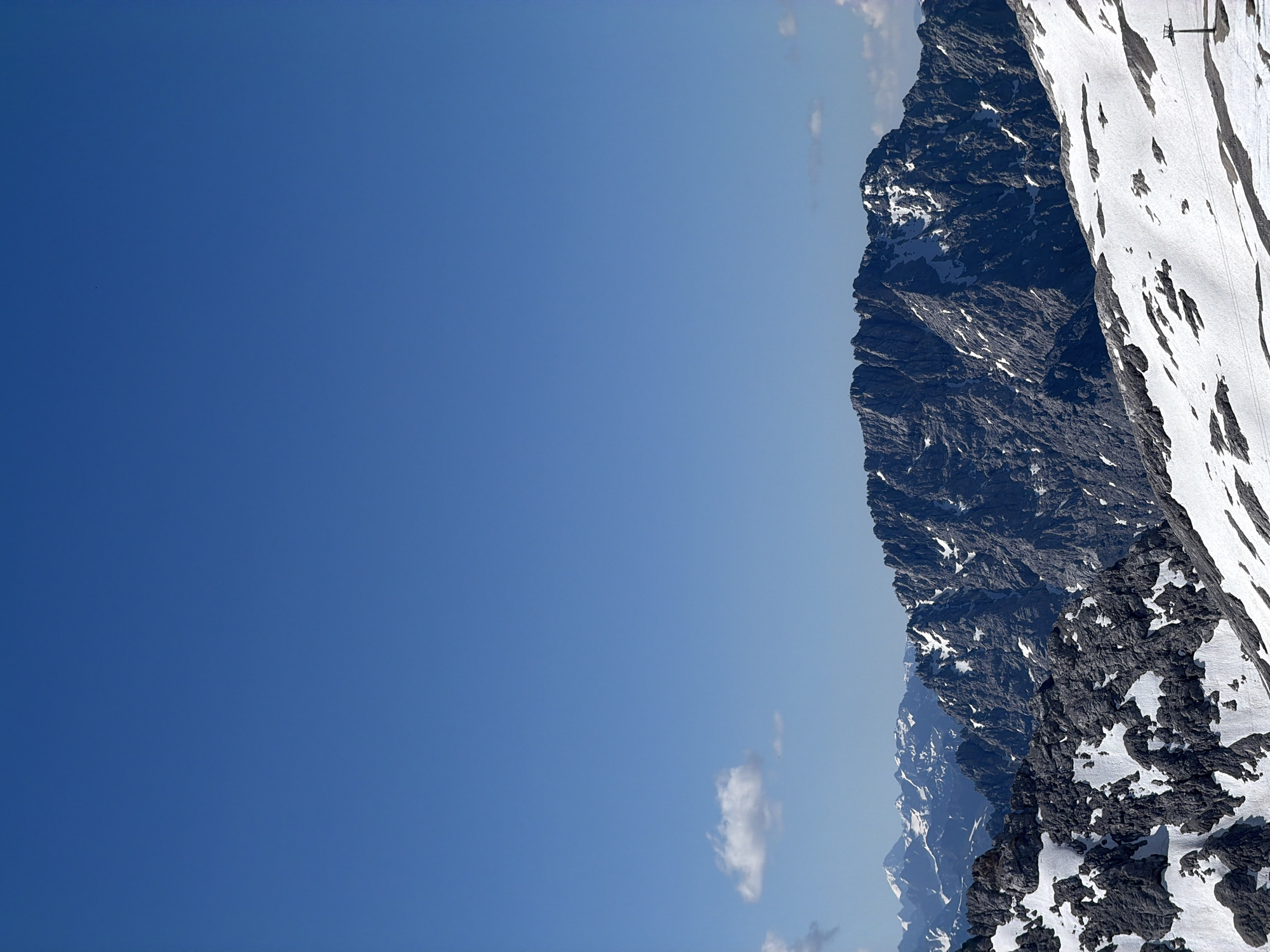 A vast, clear blue sky dominates the upper portion of the image. Below the sky, a range of rugged, snow-dusted mountains stretches across the frame. The foreground features a snowy slope with patches of dark rock exposed. In the distance, further mountain peaks are visible, also capped with snow. The overall impression is a high-altitude, natural landscape under a bright, sunny day, characteristic of alpine regions like the area around Garmisch-Partenkirchen, Germany, likely captured from a vantage point on or near the Zugspitze. There are no people or man-made structures other than what appears to be a ski lift pylon on the right side of the image.
