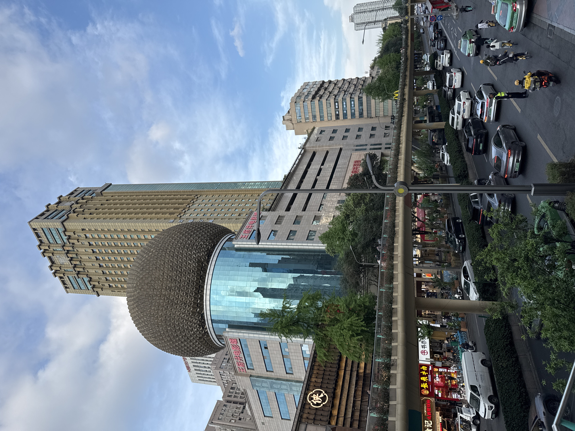 The image depicts a street scene in Jinjiang District, Chengdu, China. A tall building with a rounded, textured sphere attached to its facade dominates the left side of the frame. Adjacent to this building is another high-rise with a glass facade. Below, street-level businesses display storefront signs with Chinese characters.
A raised pedestrian walkway with several people on it is visible. On the street below, traffic is congested, including cars, motorcycles, and a traffic officer seemingly directing traffic. The sky above is partly cloudy with a mix of blue and white hues.