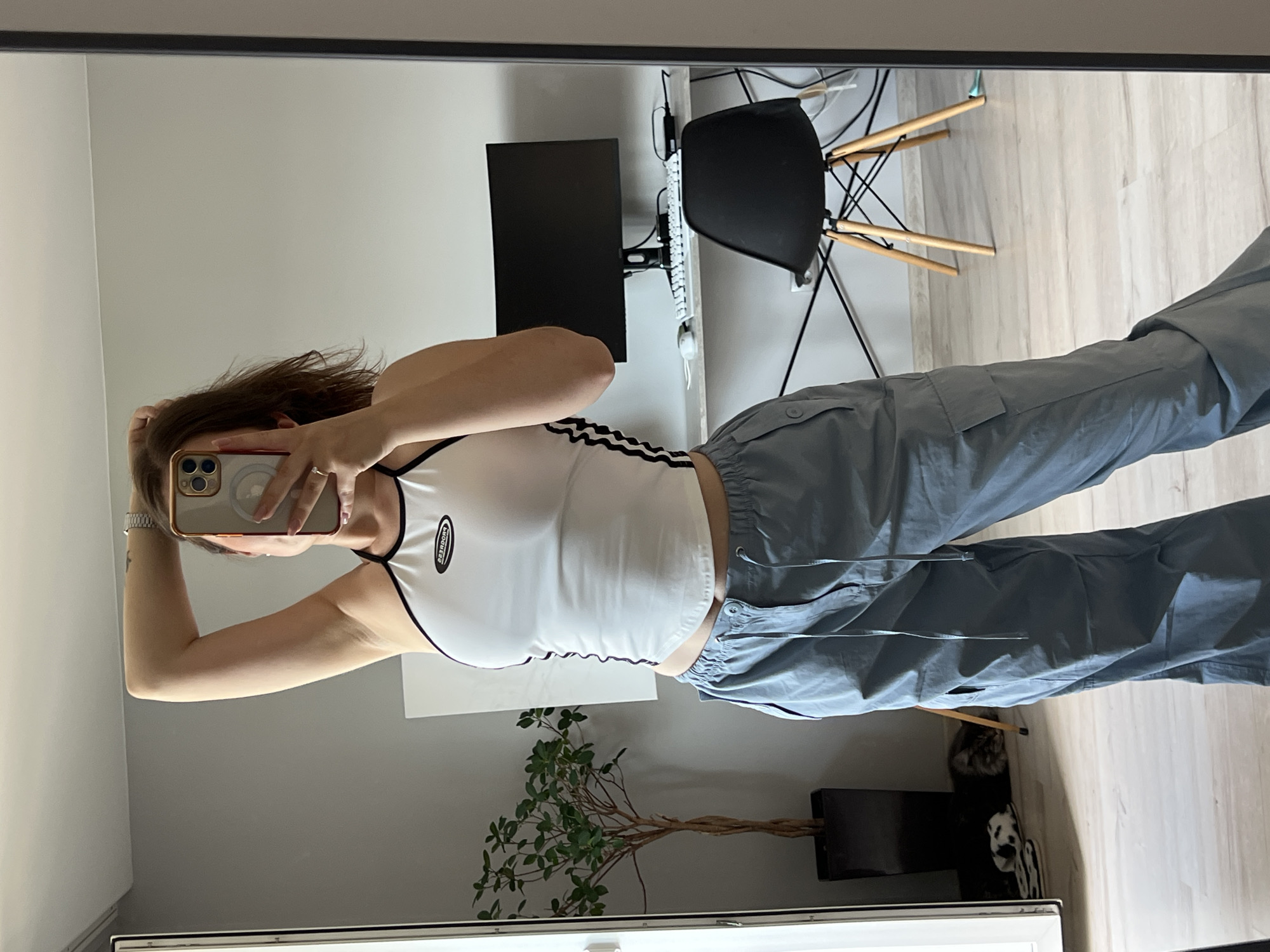 A person is taking a mirror selfie in a room. The person is wearing a white tank top with black trim and Adidas stripes, and light blue cargo pants. They have their hair pulled back and are holding a smartphone in their right hand. The background shows a desk with a computer monitor and keyboard, a black chair with wooden legs, and a plant. A cat is visible sleeping on a rug underneath the desk. The room appears to be a home office or bedroom. The lighting suggests it is daytime. There is no visible text in the image.