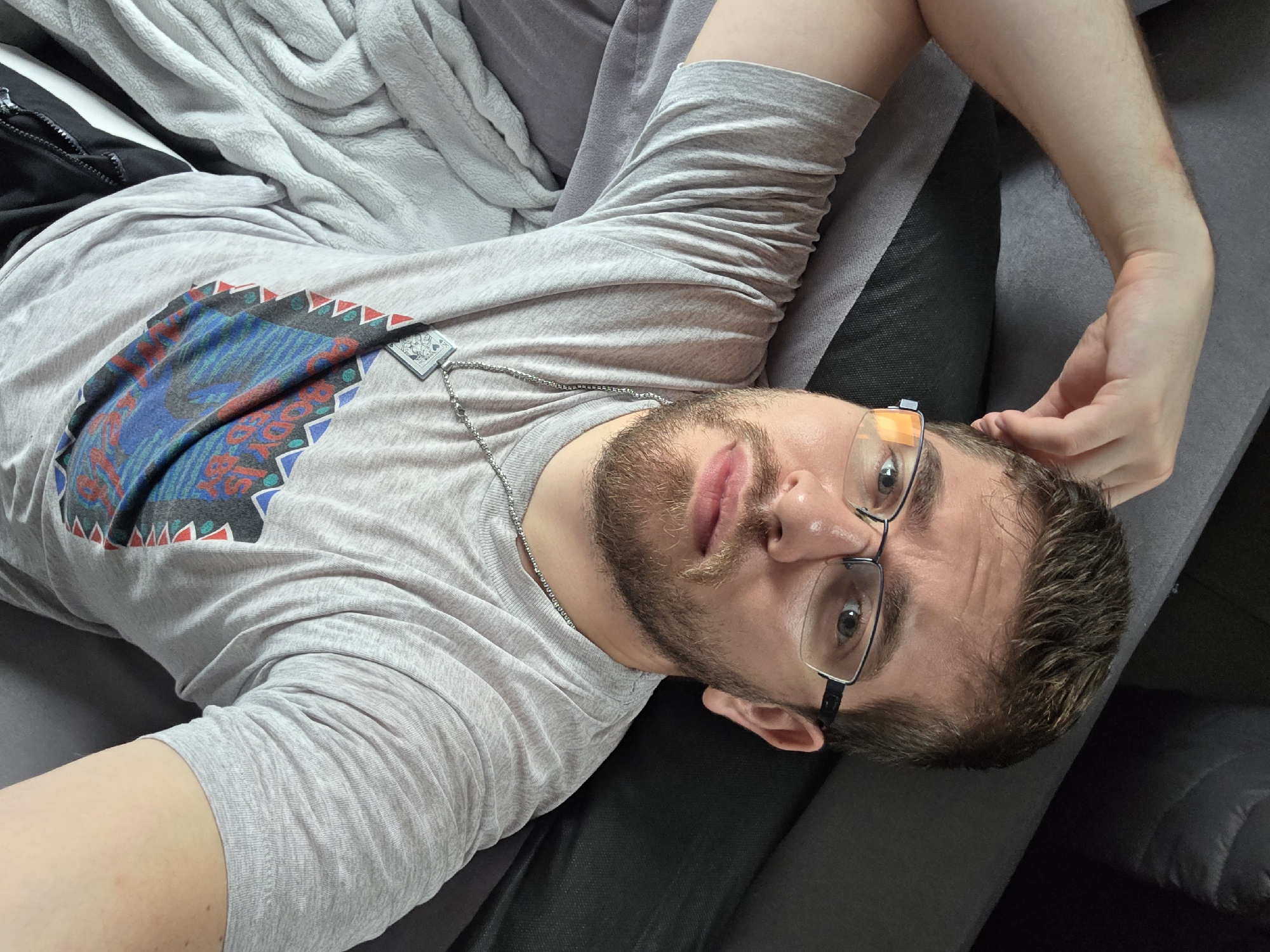 A Caucasian male with a light beard and wearing glasses is lying on a dark grey sofa. He is wearing a light grey t-shirt with a blue and red design on the chest. He has a silver chain necklace. His right arm is raised and his hand is near his head. The setting includes what appears to be a light grey blanket. There is no indication of the location being Yagodovo, Bulgaria.