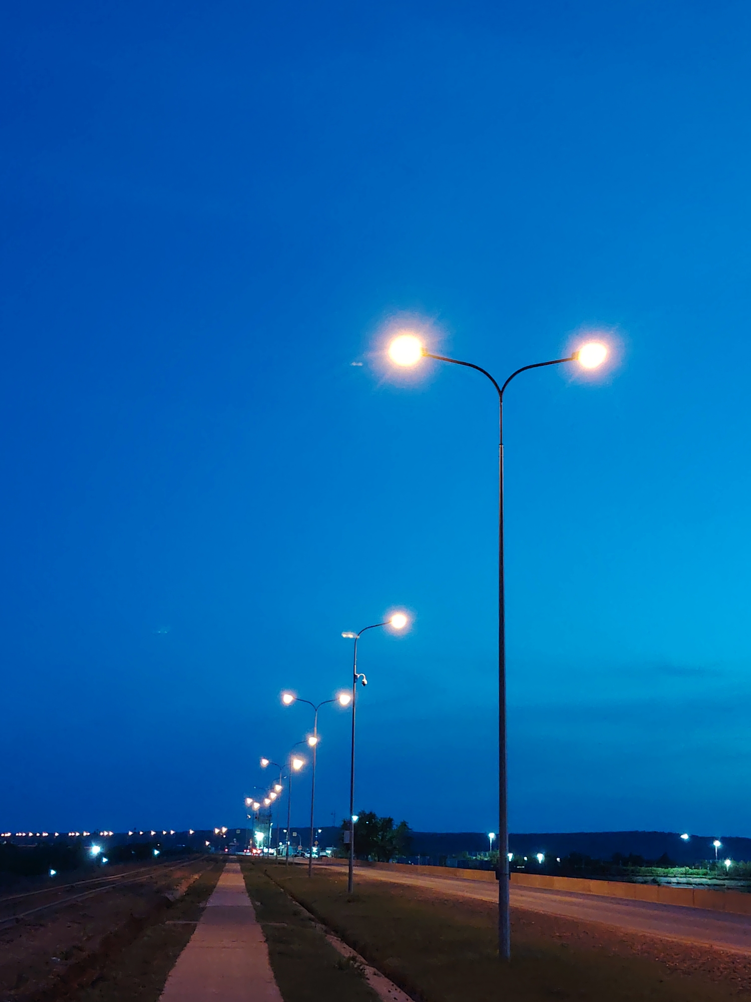 The image depicts a night scene in what appears to be a suburban or semi-rural area, possibly in Chaikovsky, Russia, given the request. A row of streetlights illuminates a paved walkway and a road, stretching into the distance. To the left of the walkway, a railway track runs parallel. Beyond the road, a line of lights suggests the presence of buildings or another road at a slightly higher elevation. The sky is a deep blue, indicating either twilight or nighttime. The scene appears quiet and devoid of human activity.