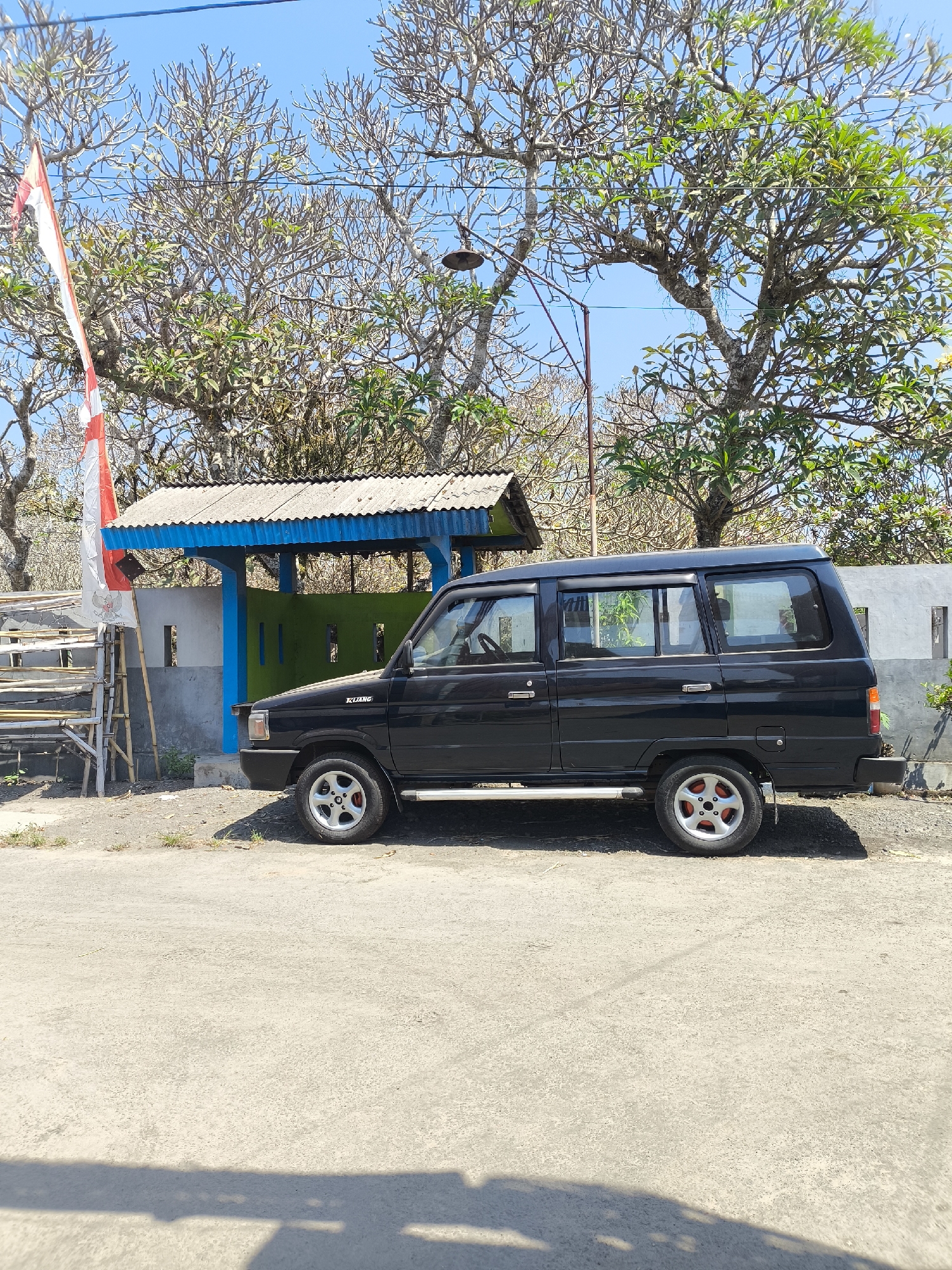 The image depicts a black Toyota Kijang parked on a paved road in Lumajang, Indonesia. The vehicle is positioned to the right of a building with a blue trim and a corrugated metal roof. A white and red Indonesian flag is visible on a bamboo pole to the left of the building. The background features several large trees with bare branches and some green foliage against a clear blue sky. The scene appears to be an outdoor setting with some dry vegetation and a light gray concrete wall behind the building. The overall impression is that of a sunny day in a somewhat rural or semi-urban area.