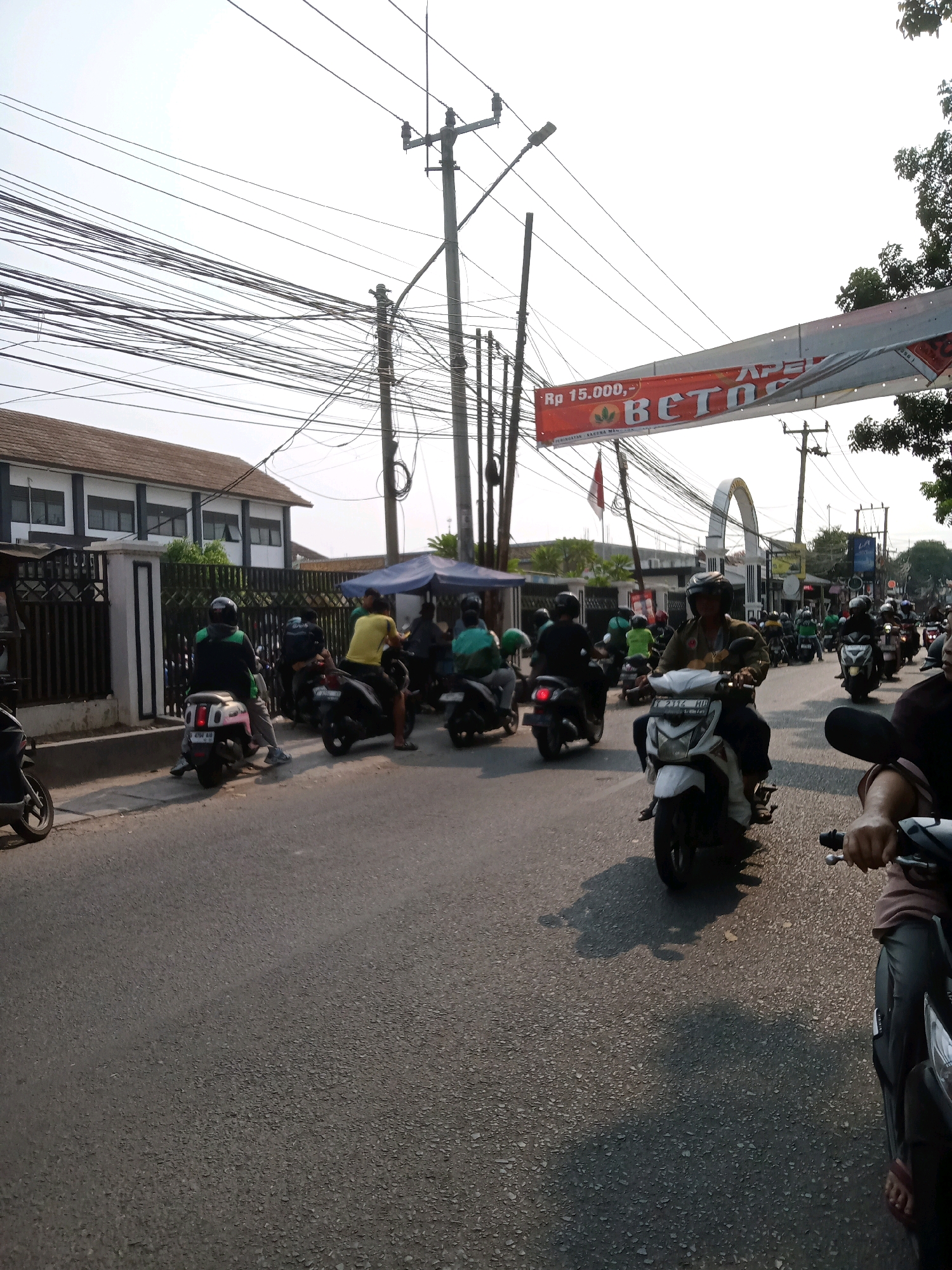The image depicts a street scene in Karawang, Indonesia, likely during daylight hours, as indicated by the bright sky and shadows. The foreground is dominated by the asphalt road, with several motorcycles visible, some in motion and others stationary. In the middle ground, a group of motorcyclists are gathered, some wearing helmets and casual attire. A building with multiple windows and a tiled roof is situated on the left side of the street, behind a black fence. A banner with red and white text, including "Rp 15.000,- BETOS", is suspended across the street, suggesting a commercial area or event. Utility poles with numerous electrical wires are prominent features, crisscrossing the sky. Trees are visible on the right edge of the frame, and an arched structure can be seen in the distance. The overall impression is a busy urban environment with moderate traffic.