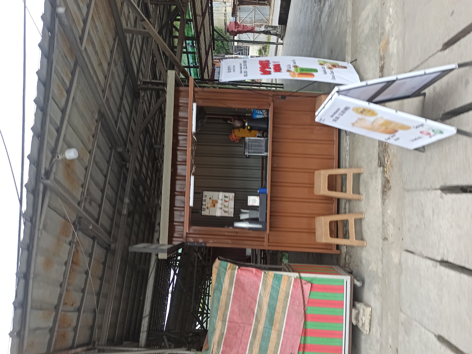 The image shows a small, open-air food stall with a brown metal counter. A menu board hangs inside the stall, displaying pictures of drinks. Two plastic stools are placed in front of the counter. To the left of the stall is another vendor cart with a striped tarp. Two A-frame signs are placed on the concrete ground in front of the stall, one displaying images of drinks and the other partially visible. The stall is located outdoors under a corrugated metal roof supported by metal beams. The surrounding environment appears to be a market or street in the City of Medan, Indonesia, with concrete pavement and some greenery visible in the background.
