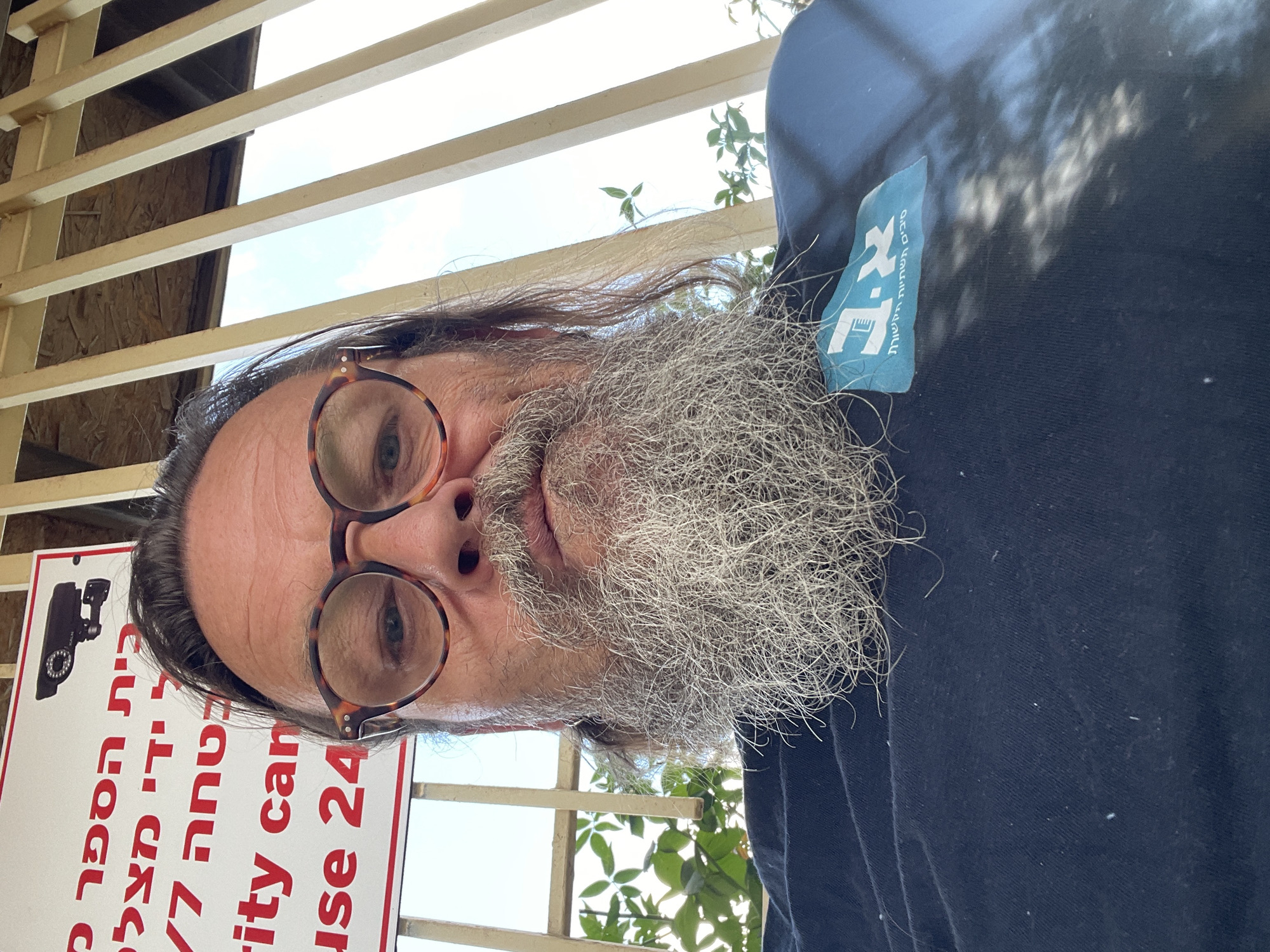 This image is a close-up selfie of an adult male with a long gray beard and glasses, taken outdoors in the city of Even Yehuda, Israel. He is wearing a dark blue t-shirt with a light blue logo on the left chest area that appears to be in Hebrew. In the background, a white sign with red lettering and an image of a security camera is visible. The sign has text in Hebrew and English. Part of the text in English reads "ity can" and "use 24". The background also shows wooden beams, foliage, and the sky.