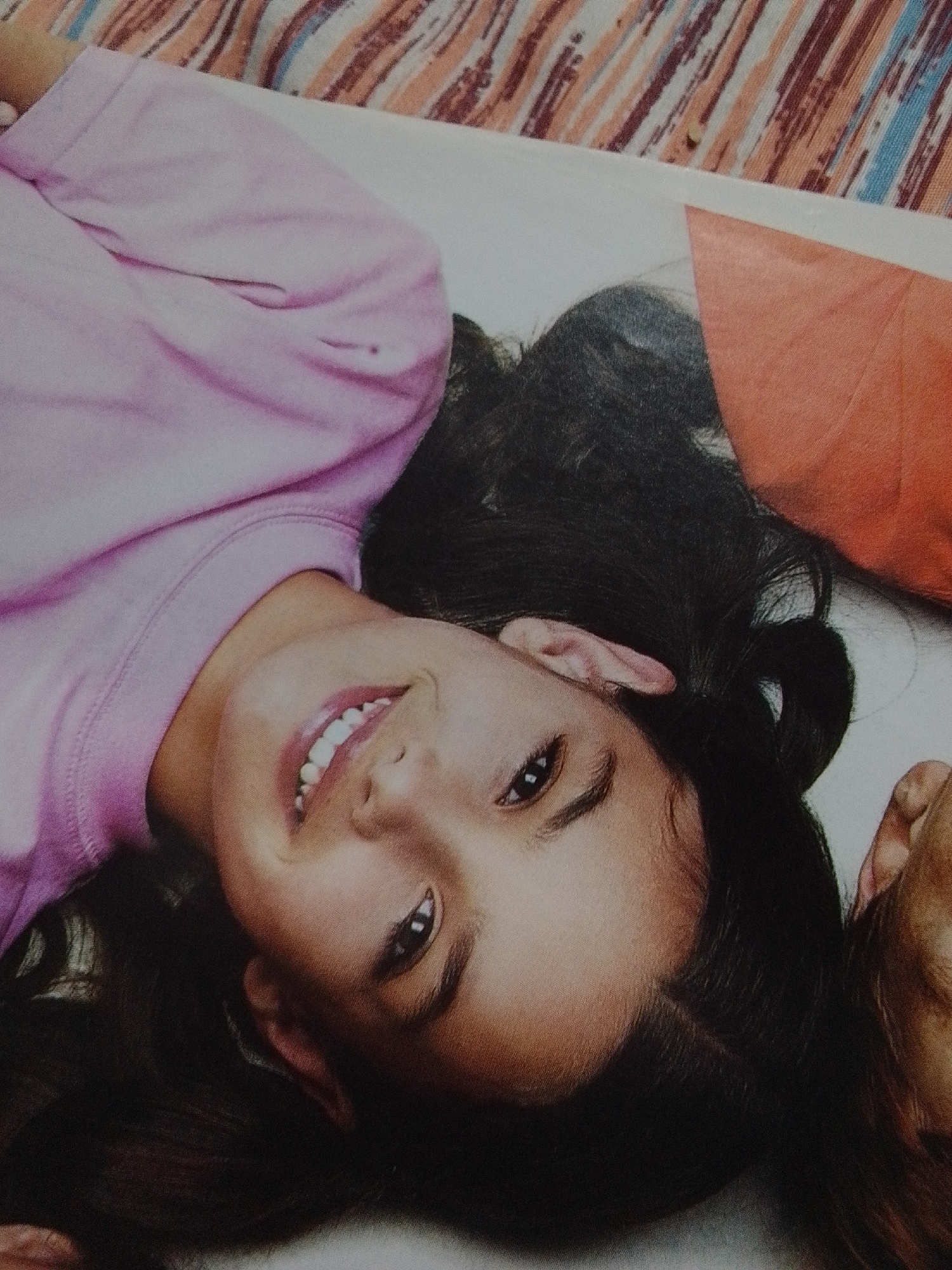 The image shows a close-up of a young girl lying down and smiling. She is wearing a pink shirt and has long, dark hair. Her head is tilted to the side, and she is looking directly at the camera. The background is a colorful, striped fabric. There are no visible objects, text, or clear cues about the time of day, weather, or specific location. The girl's smile suggests a happy or playful mood. The scene appears to be indoors or in a controlled environment, given the fabric background.