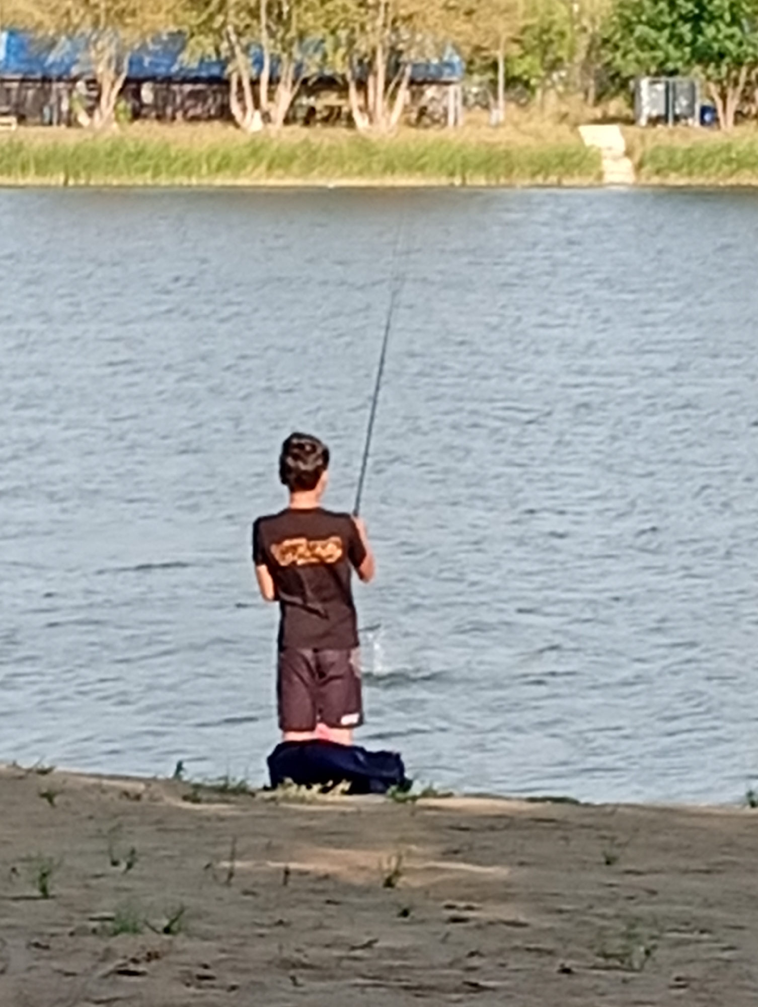 A lone male is fishing from the shore of a body of water. He is wearing a black t-shirt and dark shorts. He is holding a fishing rod, with the line cast into the water. A dark blue bag or bundle is on the ground behind him. The background shows a shoreline with some vegetation and trees, as well as some buildings and structures further back. The water appears calm, with ripples on the surface. The lighting suggests it is daytime, likely late afternoon given the shadows. The scene is likely a public park or recreational area along a river or lake in Kostanay, Kazakhstan.