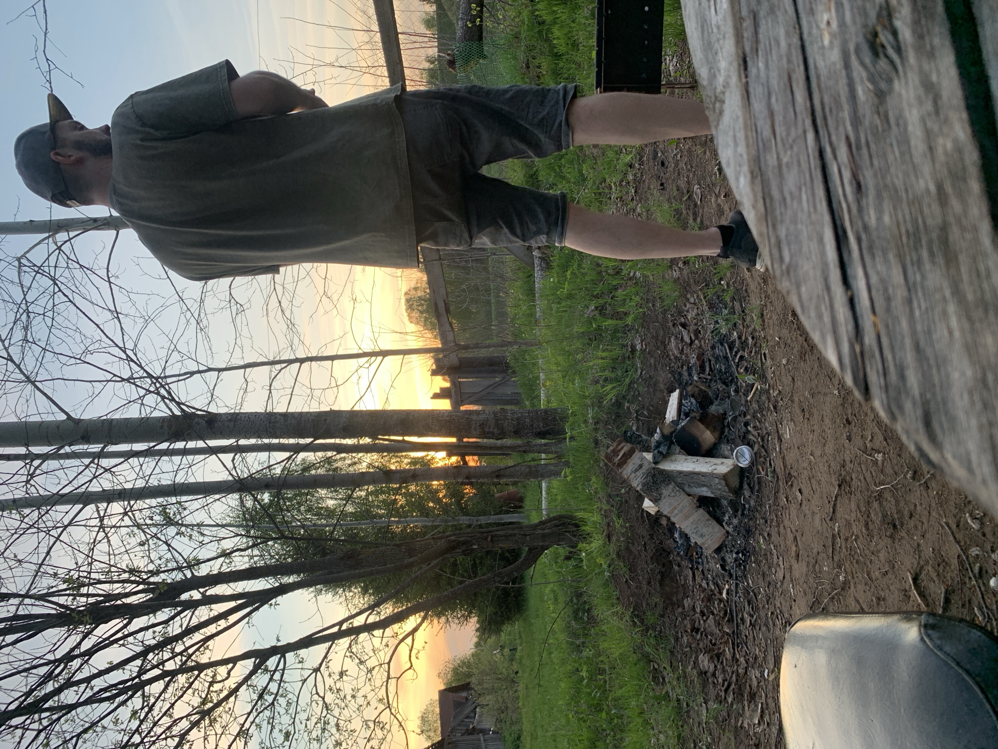A man wearing a cap, t-shirt, and shorts stands outdoors near a wood pile and a wooden structure. The environment includes trees, grass, and what appears to be a sunset in the background. The location appears to be rural, with buildings visible in the distance. The city of Zasechnyi, Russia, is not directly confirmed but presumed, without additional information.