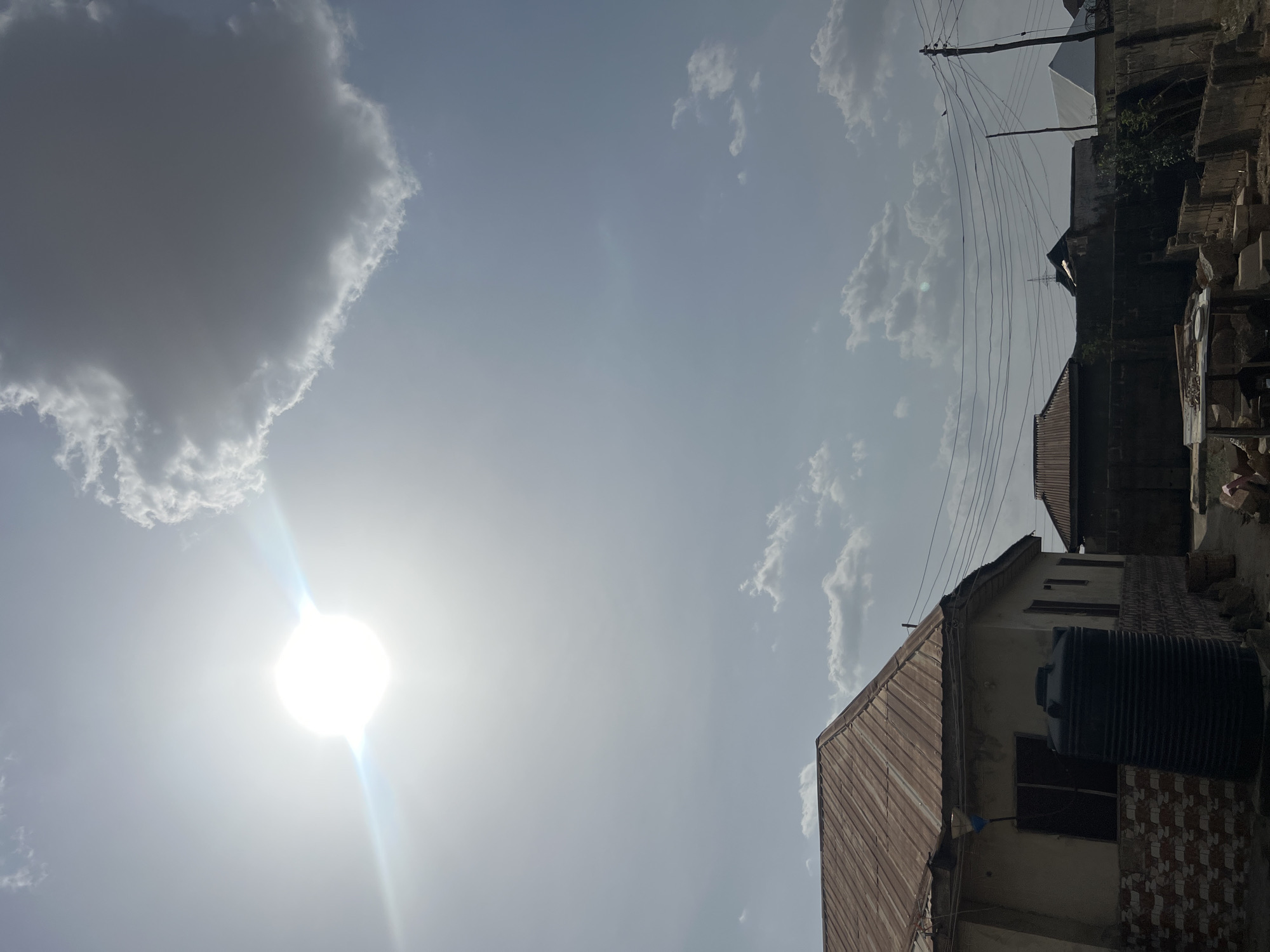 The image shows a daytime scene in Minna, Nigeria. The sky is mostly clear with some cumulus clouds and the sun shining brightly. In the foreground, there is a building with a corrugated metal roof and a large, black water tank. Power lines and a utility pole are visible above the building. Other buildings are visible in the background, suggesting a residential area.